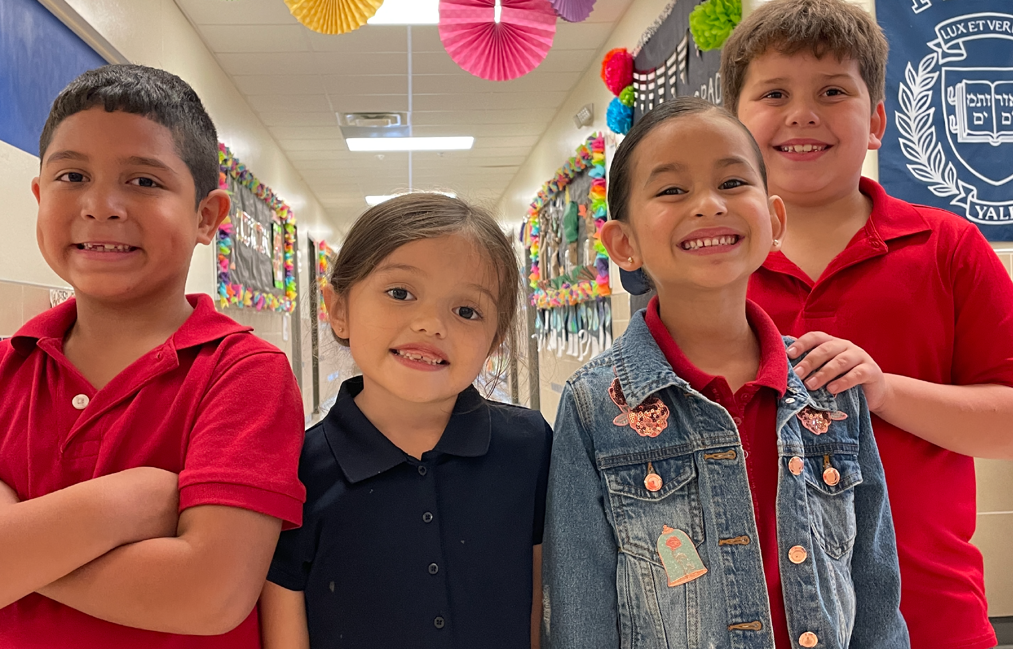 Four smiling children wearing red and blue shirts stand in a colorful, decorated school hallway.