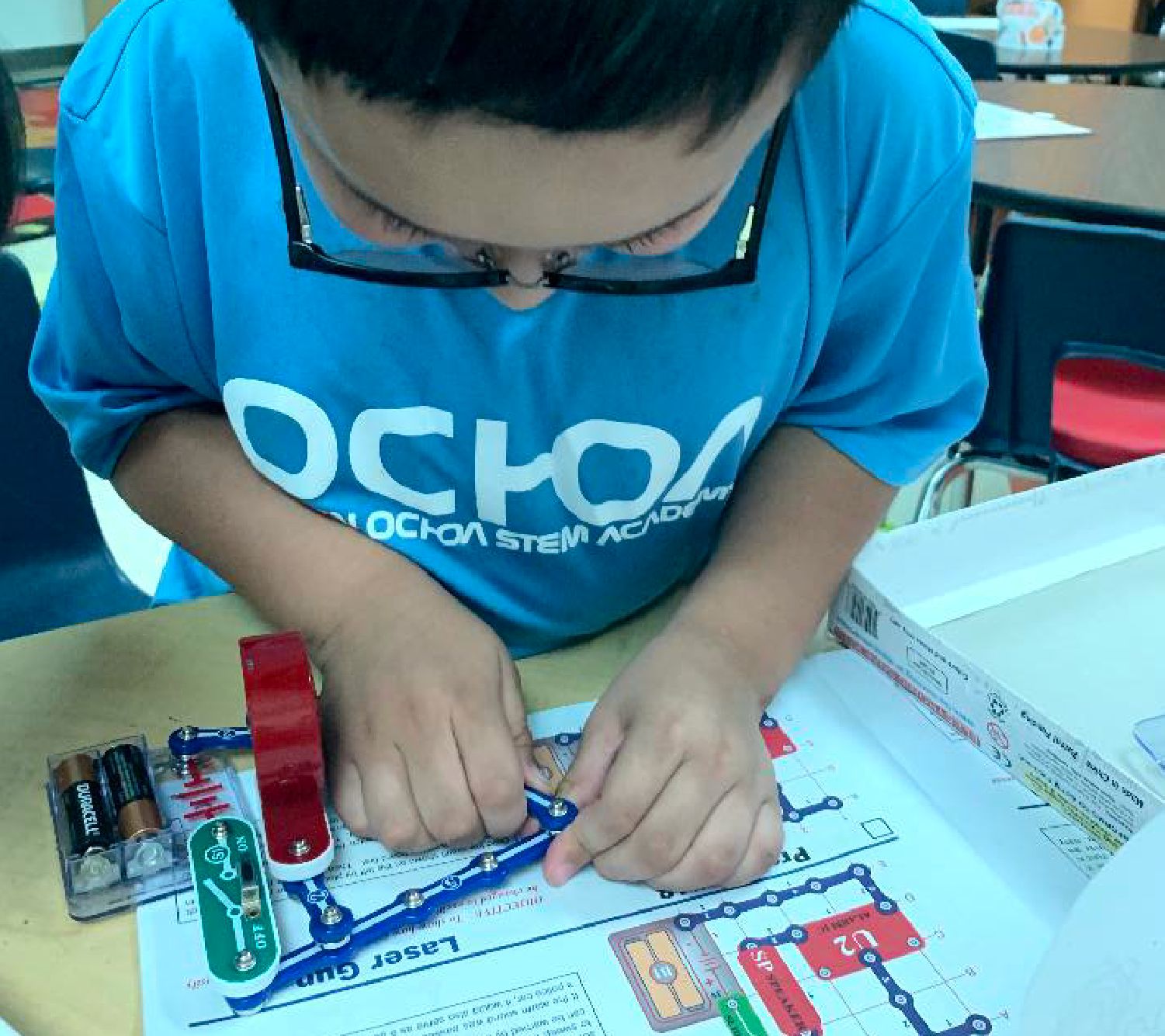 The image shows a person wearing a blue shirt with the text "OCOA" on it, sitting at a table and working on something with various tools and materials in front of them.