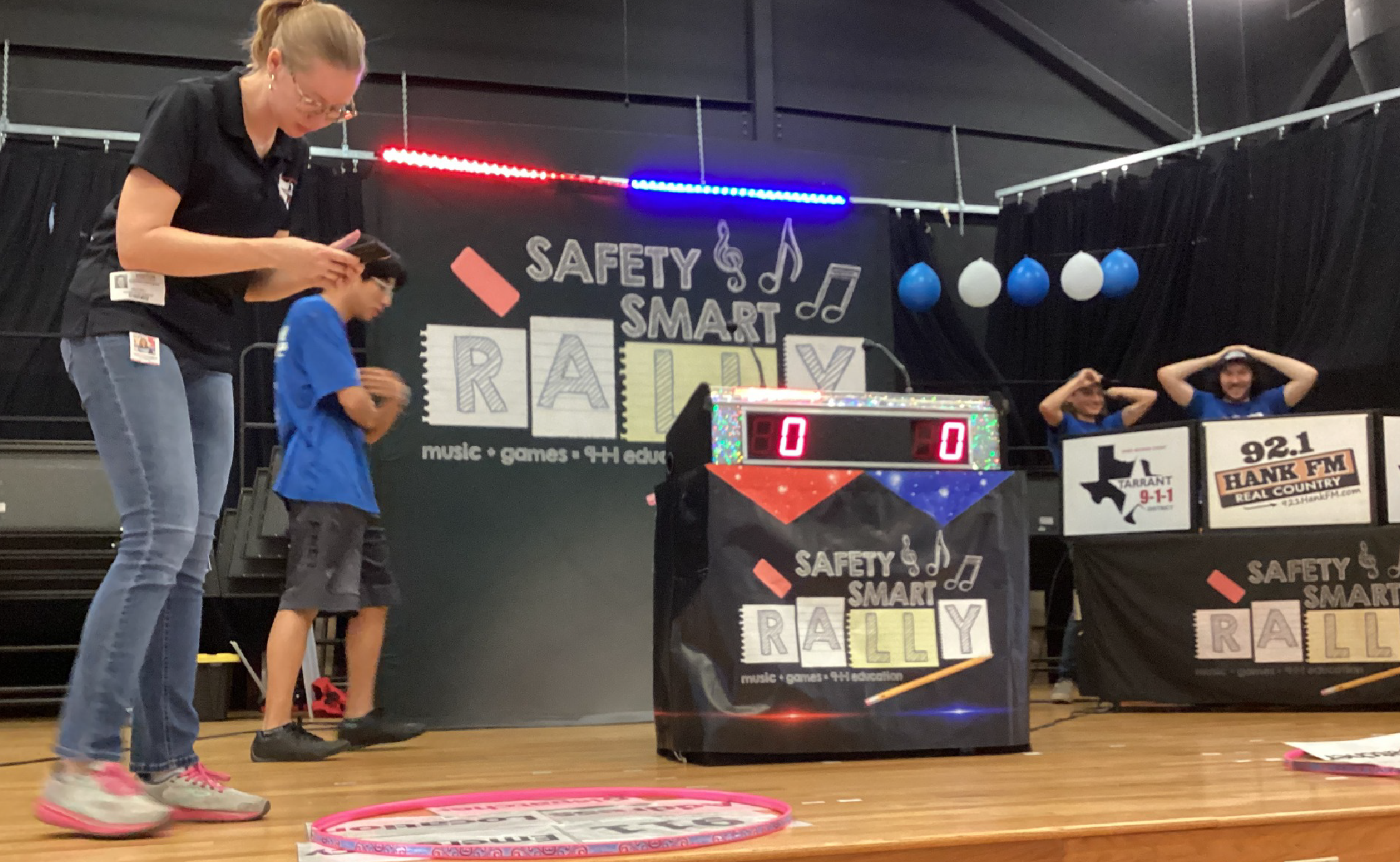 The image shows a person standing in front of a large display board with various safety-related information and equipment, including a digital countdown timer, balloons, and other safety-related items.