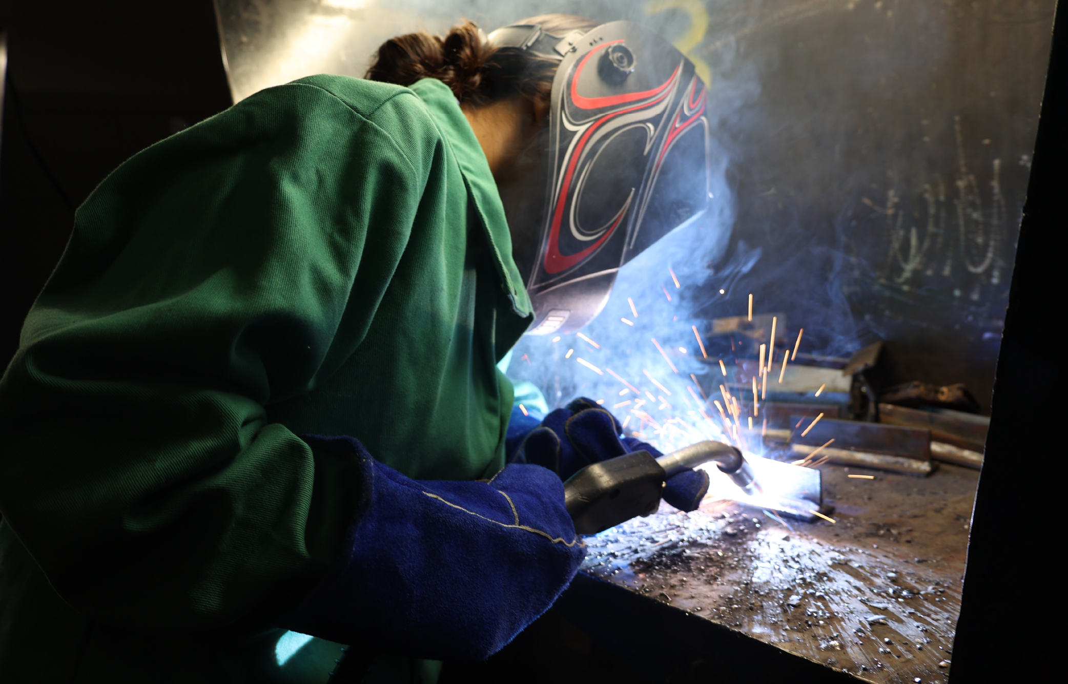 A person in a green jacket and blue gloves is using a welding tool, creating sparks in a dimly lit industrial setting with graffiti-covered walls in the background.
