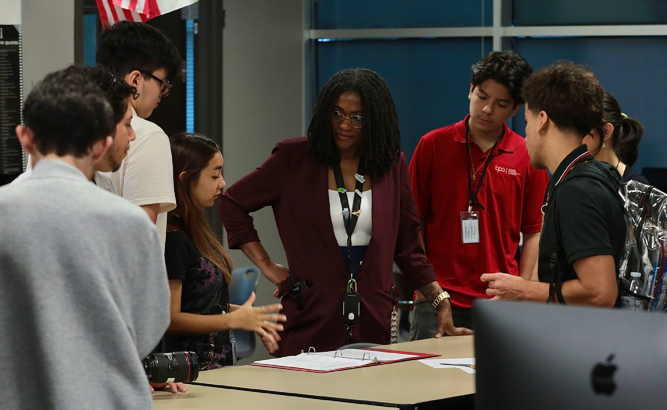 A group of people, some wearing red shirts, are gathered around a table and appear to be engaged in discussion or collaboration.