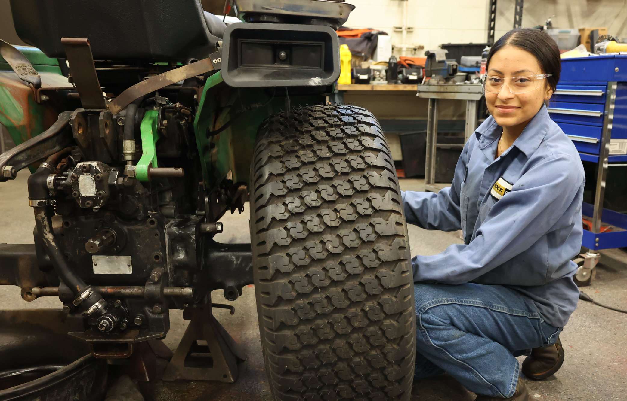 A female mechanic in a blue uniform is inspecting the tire of a large agricultural vehicle in a workshop setting with various tools and equipment visible in the background.