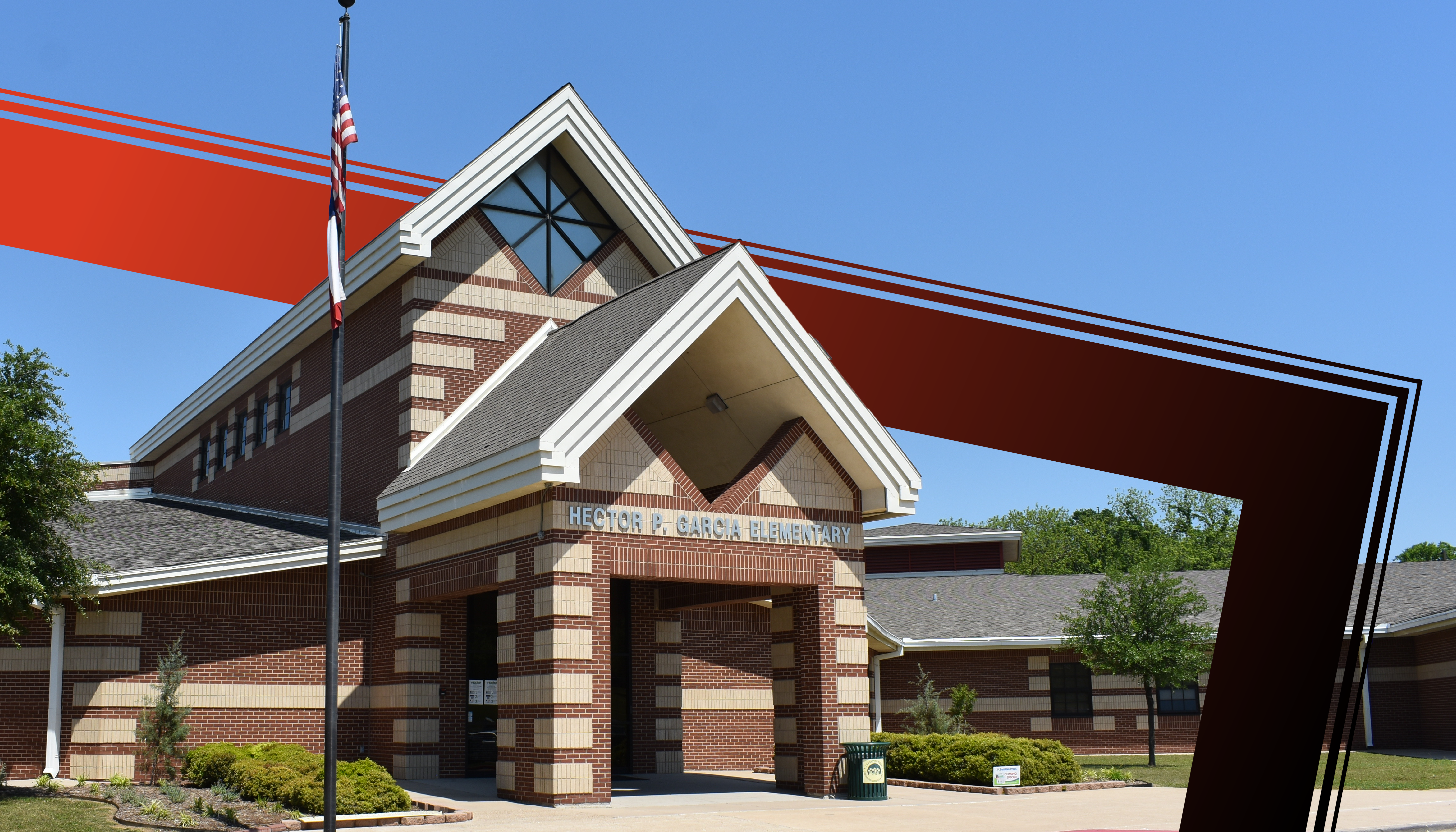 A two-story brick building with a triangular roof and large windows, surrounded by trees and greenery, against a clear blue sky.