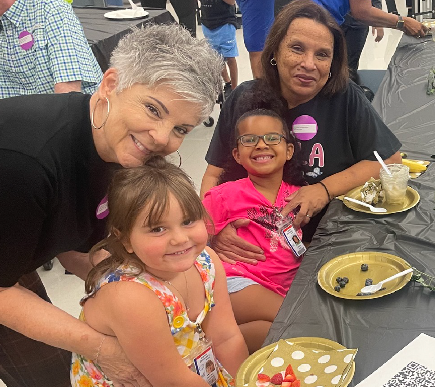 The image shows three women, two young girls, and a table with food in front of them, suggesting a casual gathering or event.