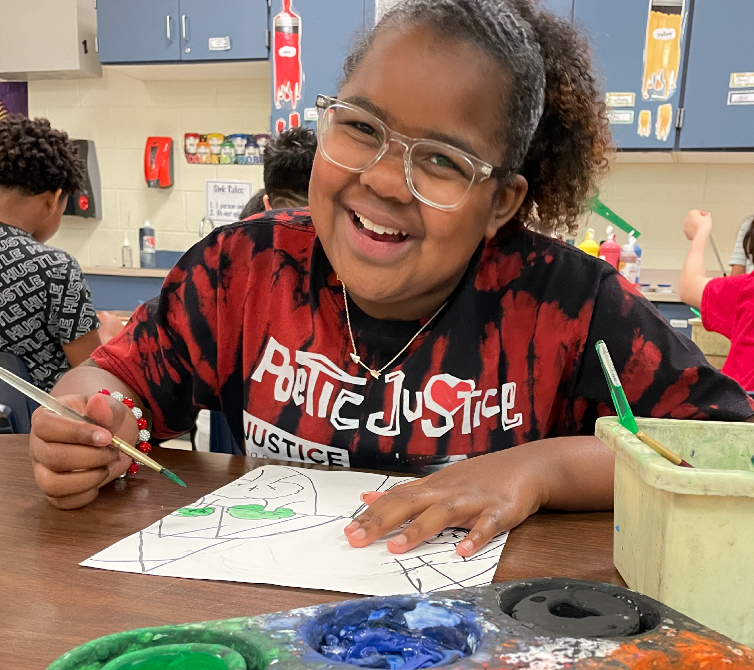 A smiling young person wearing a red and black patterned shirt and glasses is sitting at a desk, drawing on a piece of paper in a classroom setting with various educational materials visible in the background.