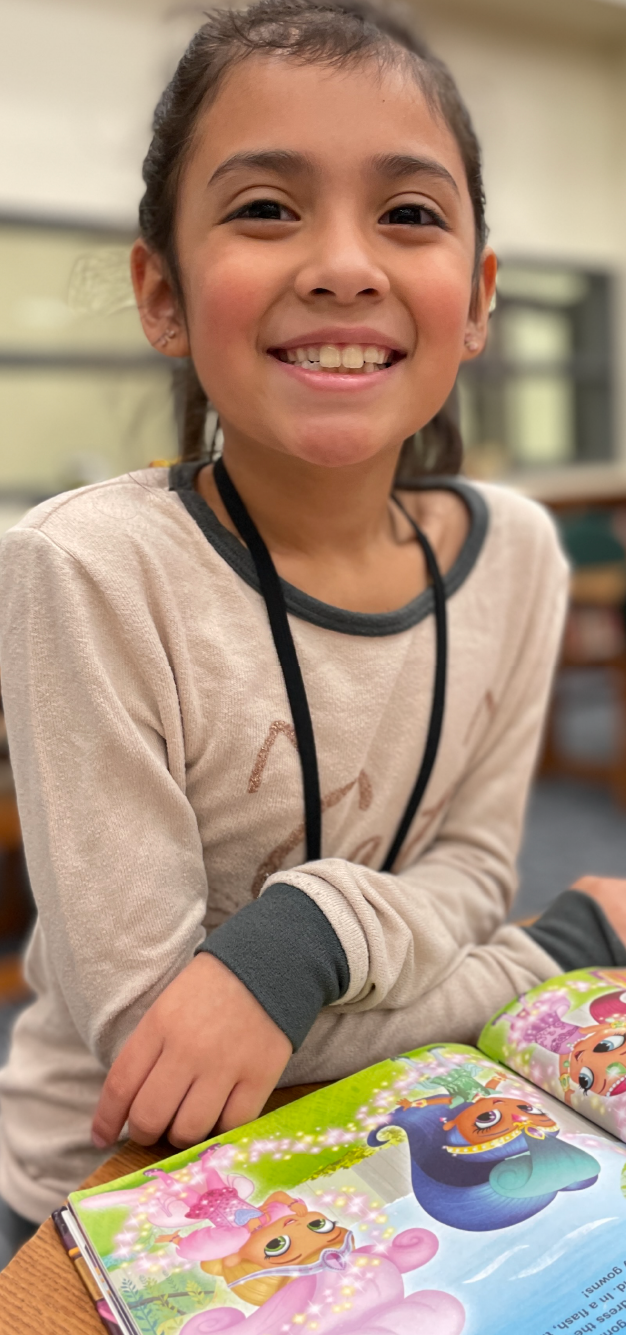 A young girl with a warm smile is sitting at a table, surrounded by colorful books and toys.