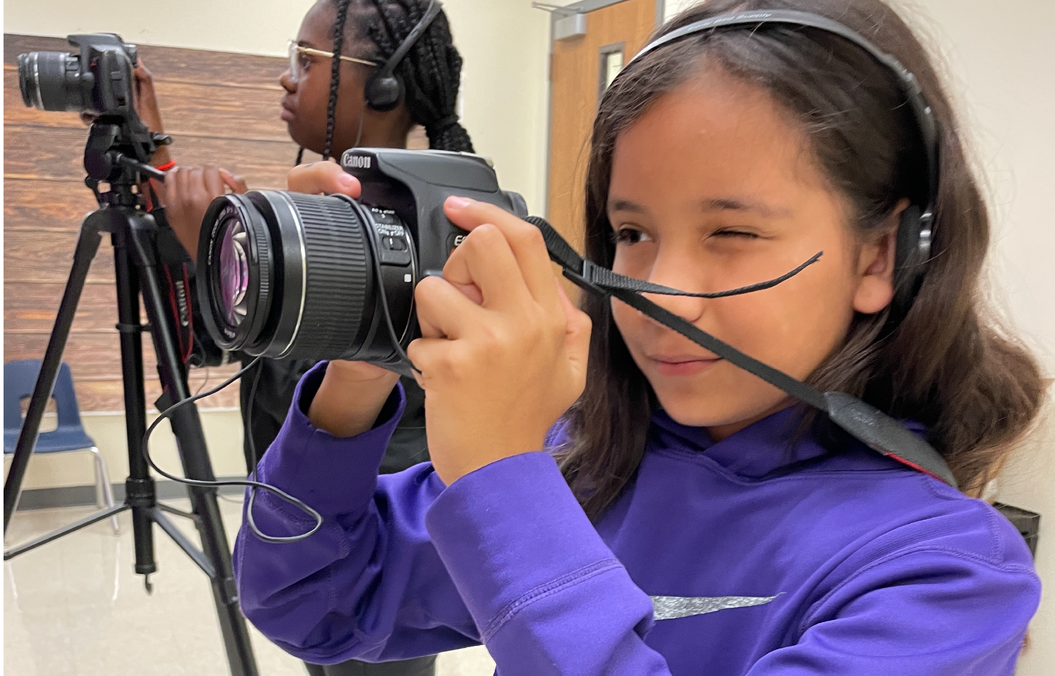 A young girl in a purple sweater is holding a camera and appears to be taking a photograph, with a tripod and other photographic equipment visible in the background.
