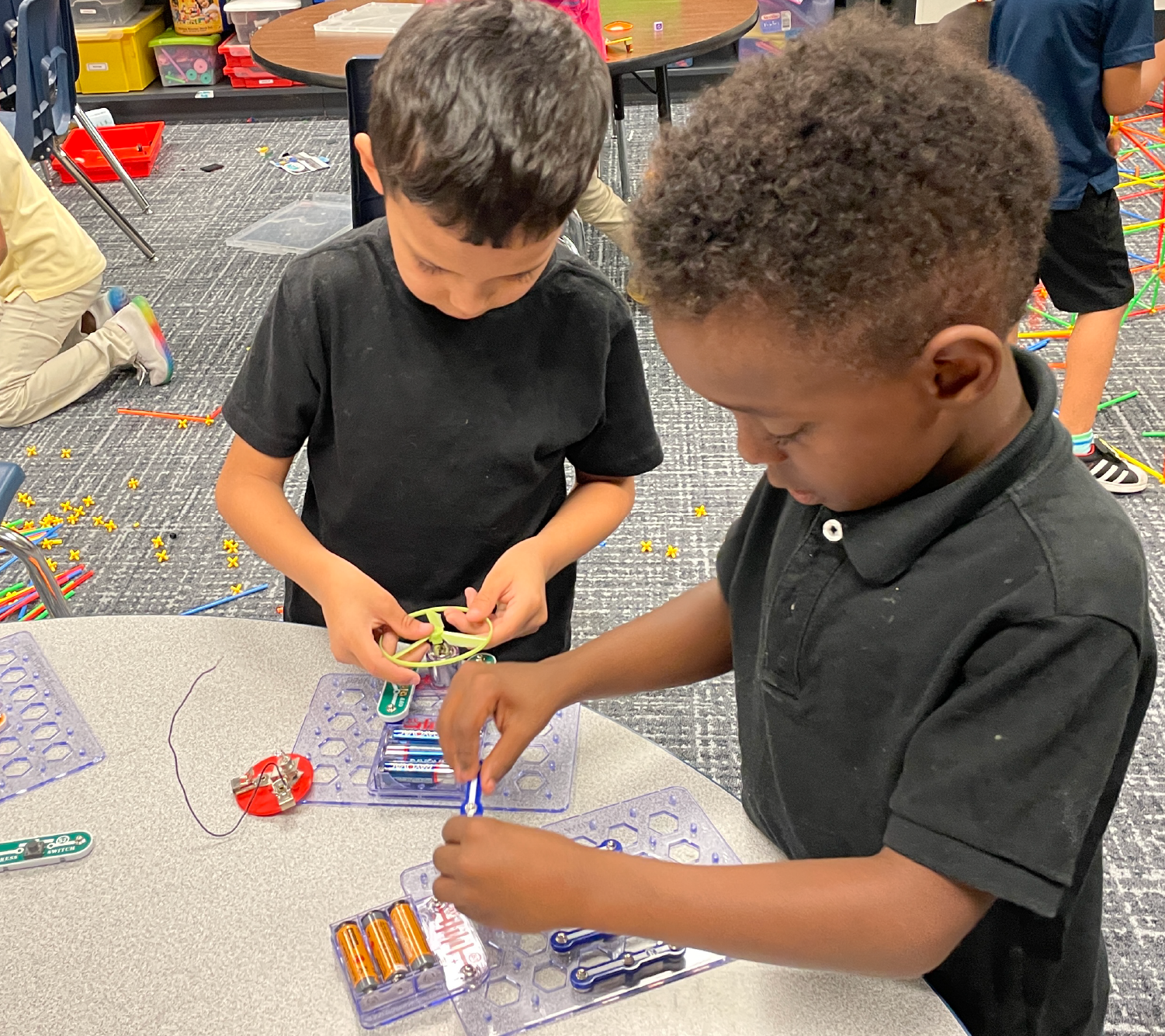 Two young boys, one with curly hair and the other with short hair, are sitting at a table and engaged in a hands-on activity, surrounded by various school supplies and materials in the background.
