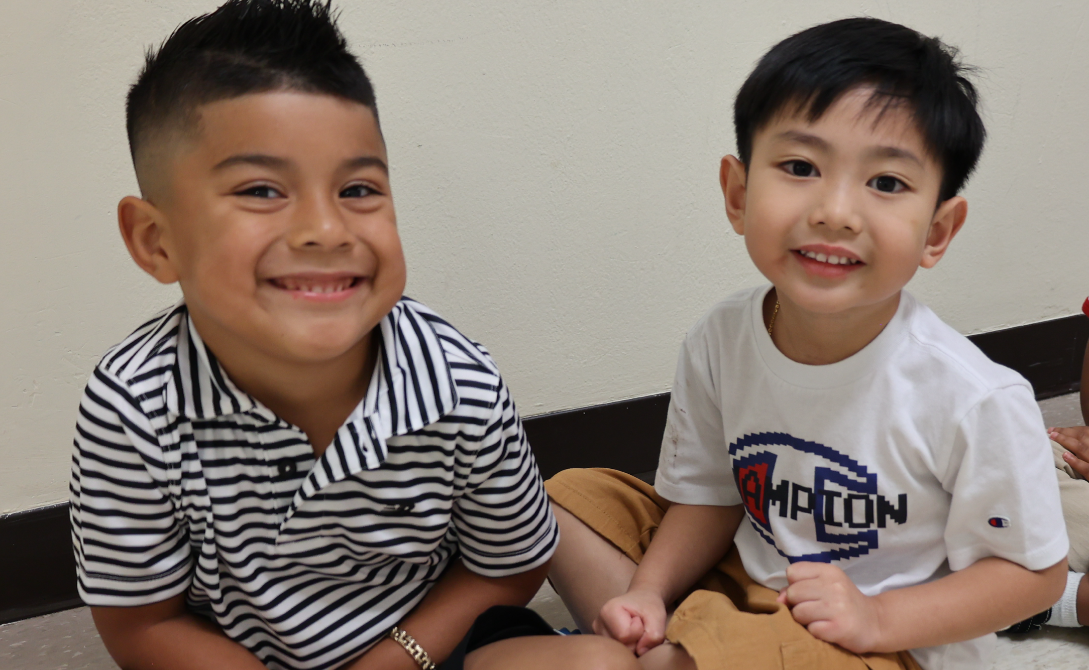 Two young boys with cheerful expressions, one wearing a striped shirt and the other a white t-shirt, sitting together in what appears to be a living room or home setting.