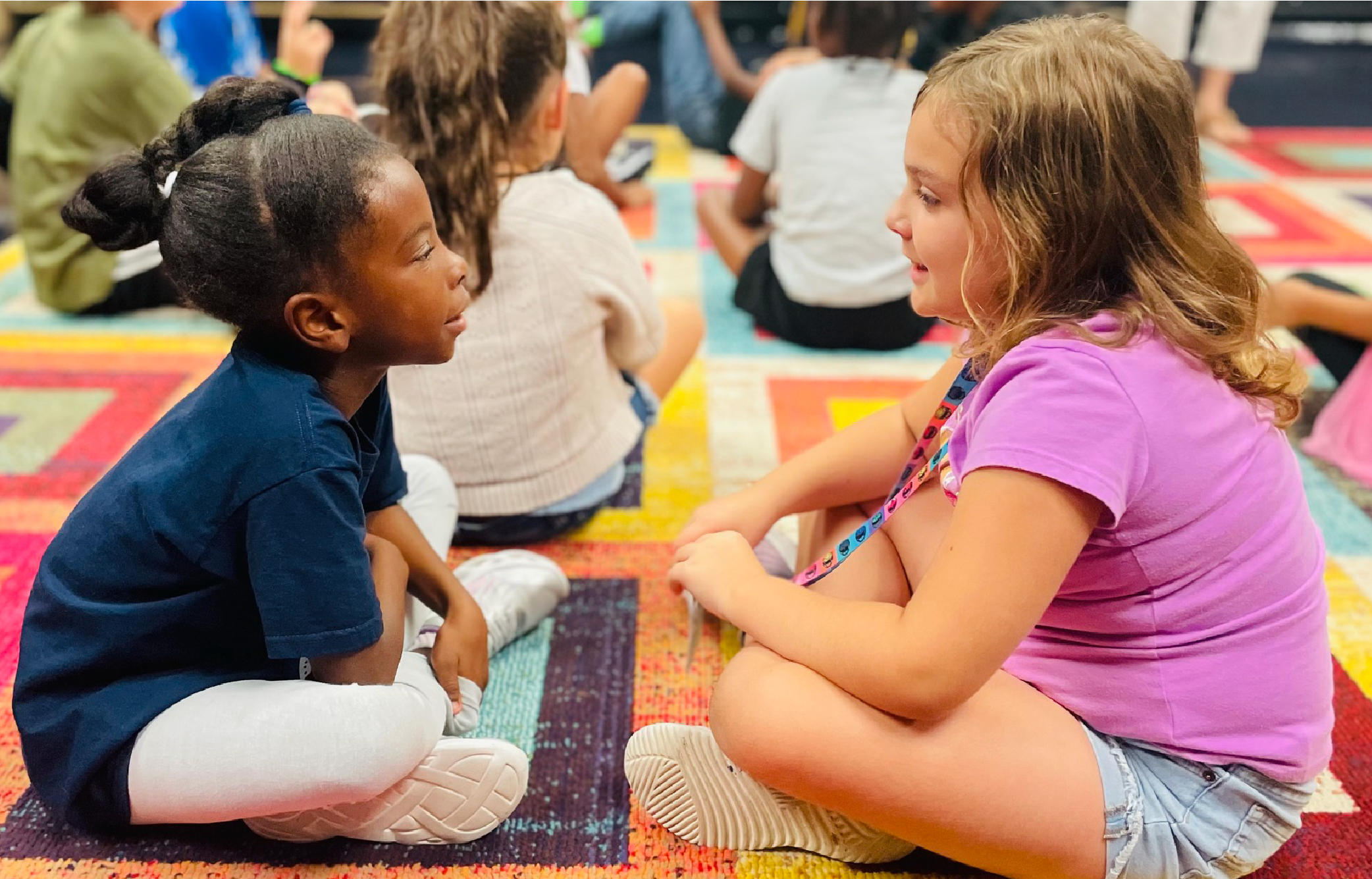 Two young girls, one with dark hair and one with light hair, are sitting on the floor and engaging in conversation. The background appears to be a colorful, patterned rug or mat, suggesting an indoor setting.