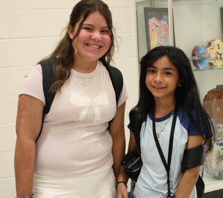 The image shows two young women, one with long brown hair and the other with long dark hair, standing together and smiling in what appears to be a school or office setting, with colorful artwork visible in the background.