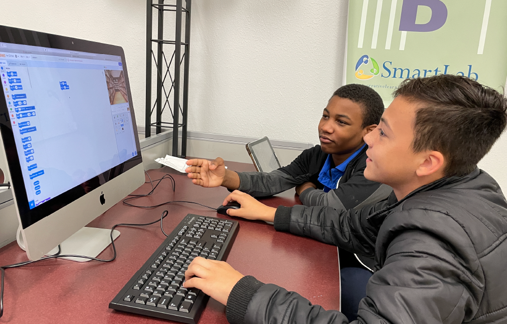 Two individuals, a man and a boy, are seated at a computer workstation, intently focused on the screen in front of them. The background features a sign that reads "Smart Lab", suggesting an educational or technology-focused setting.