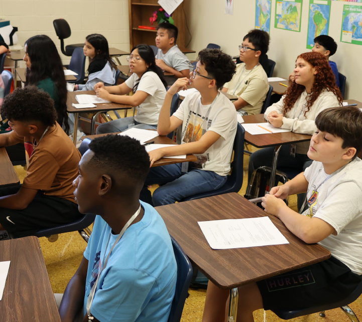 A classroom setting with students seated at desks, engaged in what appears to be a lesson or discussion, with various educational materials and posters visible in the background.