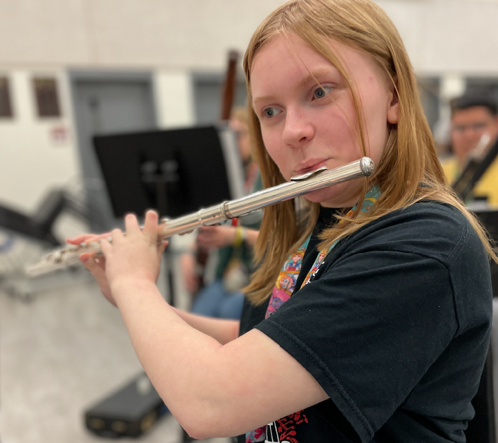 A young woman with long blonde hair is playing a silver flute in what appears to be a music classroom or rehearsal space, with various musical instruments and equipment visible in the background.