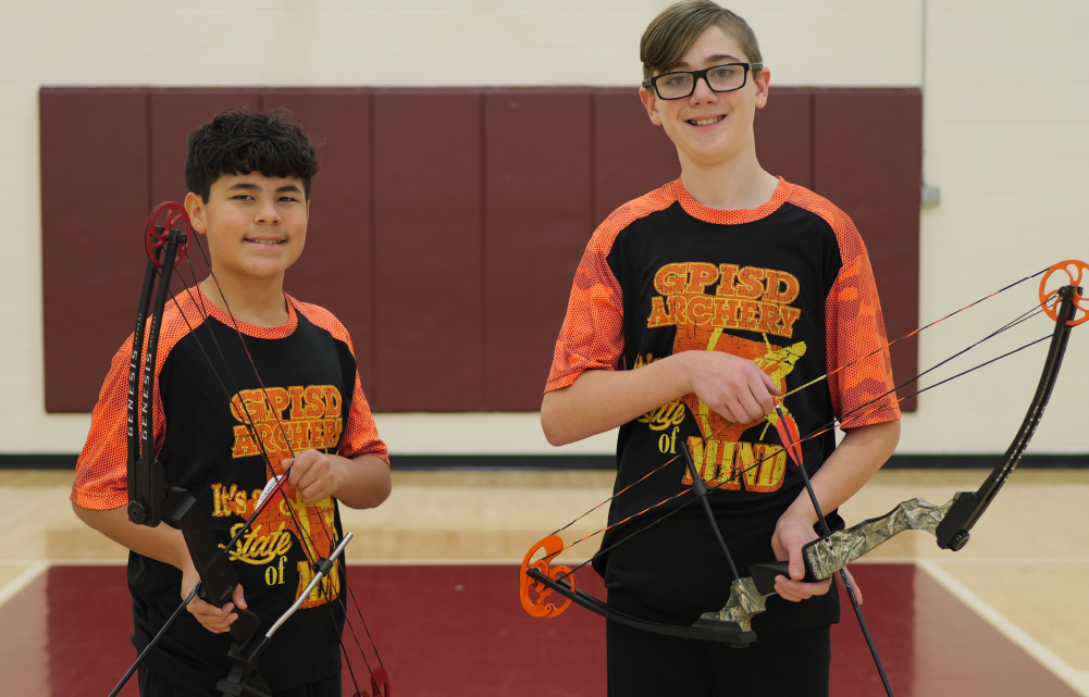 Two young individuals wearing matching orange and black jerseys stand in a gymnasium setting, smiling and holding what appear to be gaming controllers.
