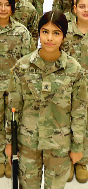 A young woman in military uniform stands among a group of soldiers in a camouflage setting.