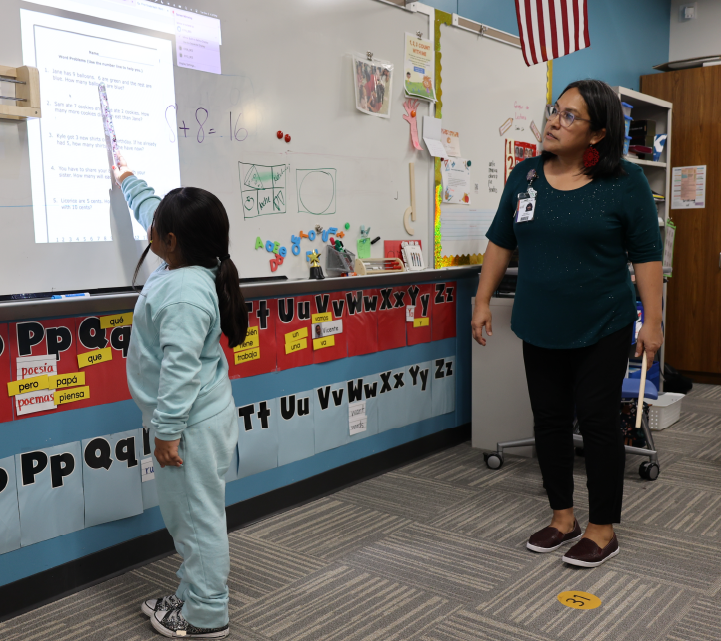 The image shows a classroom setting with a teacher and a student interacting in front of a colorful display board.