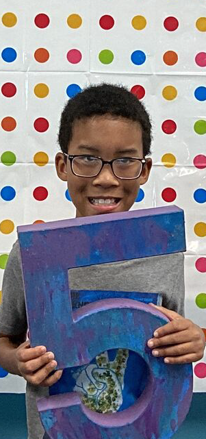 A young boy with glasses is smiling and holding up a colorful, abstract painting in front of a patterned background of multicolored dots.