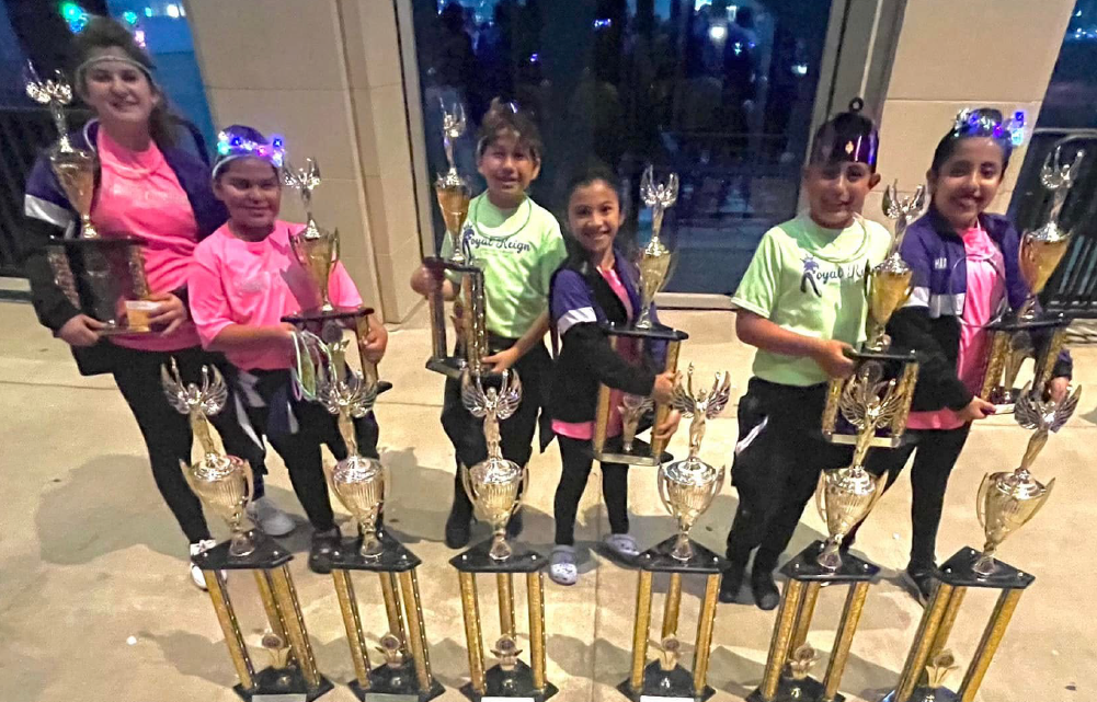 A group of young girls, dressed in colorful athletic wear, are standing together and holding various trophies or awards in a dimly lit room with decorative lighting fixtures.
