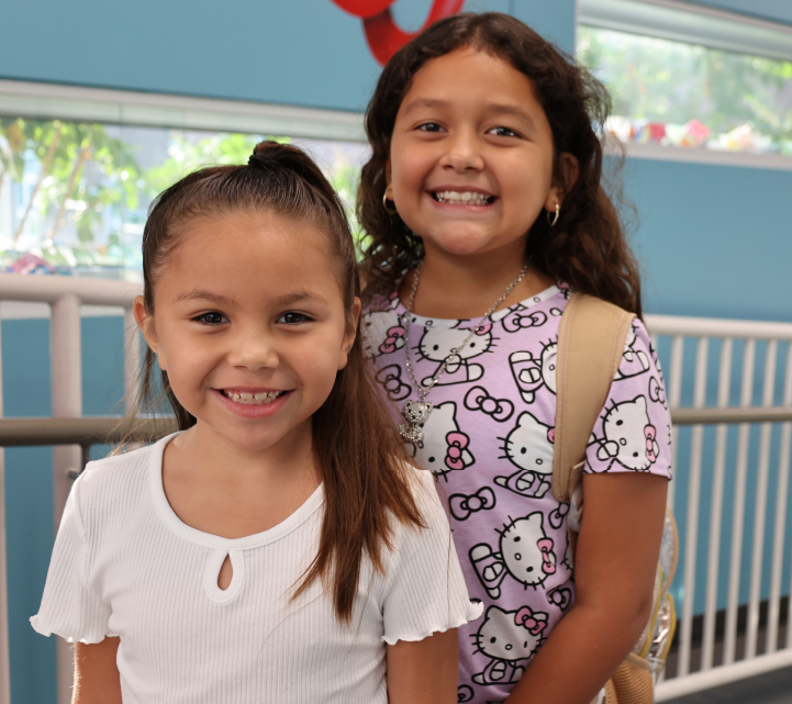 Two young girls with smiling faces, one wearing a white shirt and the other a patterned dress, standing in front of a blue wall with decorations.