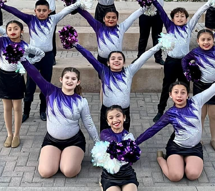 A group of young people in cheerleading uniforms, with purple and white colors, are posing together outdoors on a paved surface.