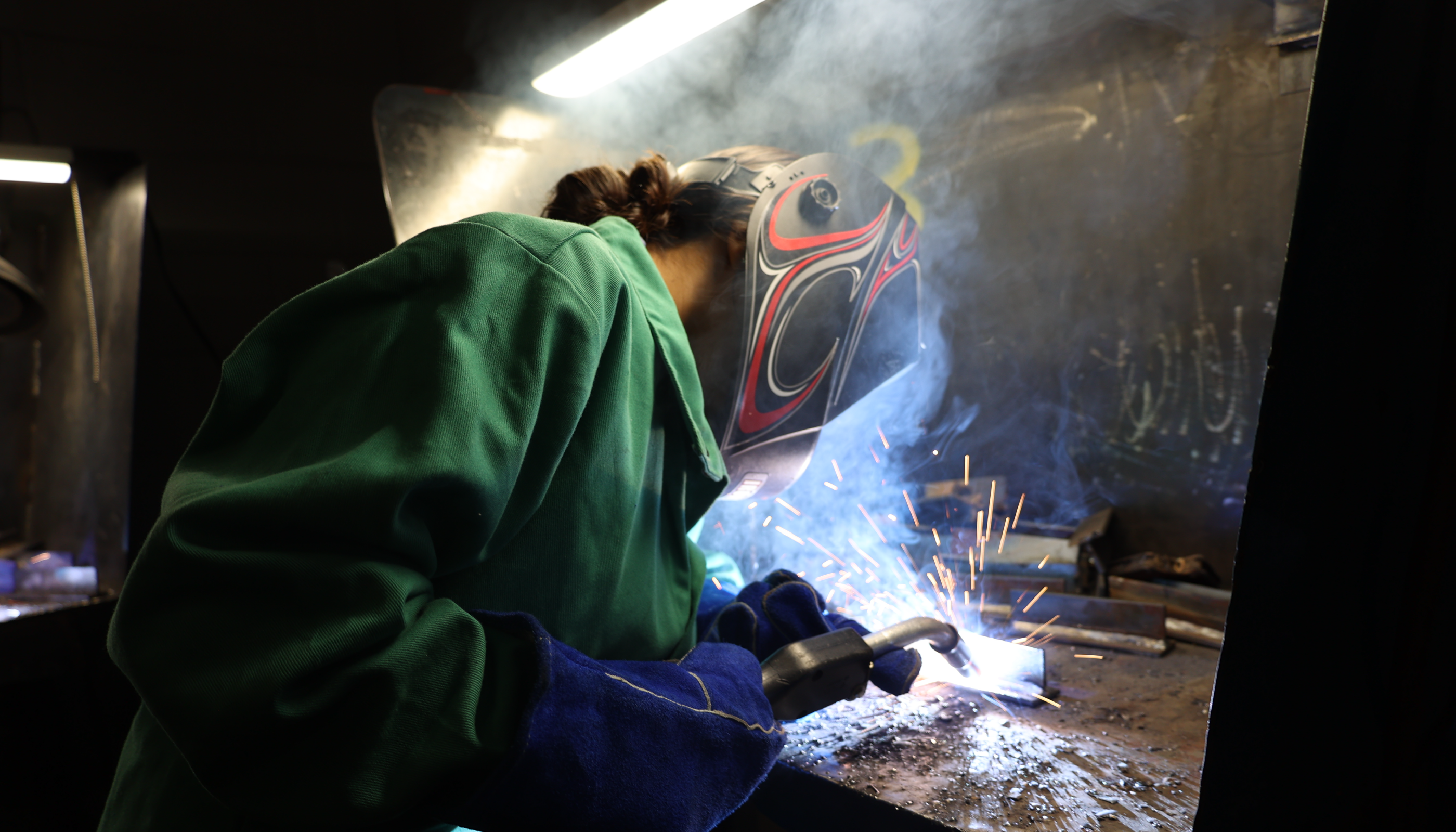 A person in a green protective suit is using a welding tool, creating sparks and smoke in a dimly lit industrial setting.