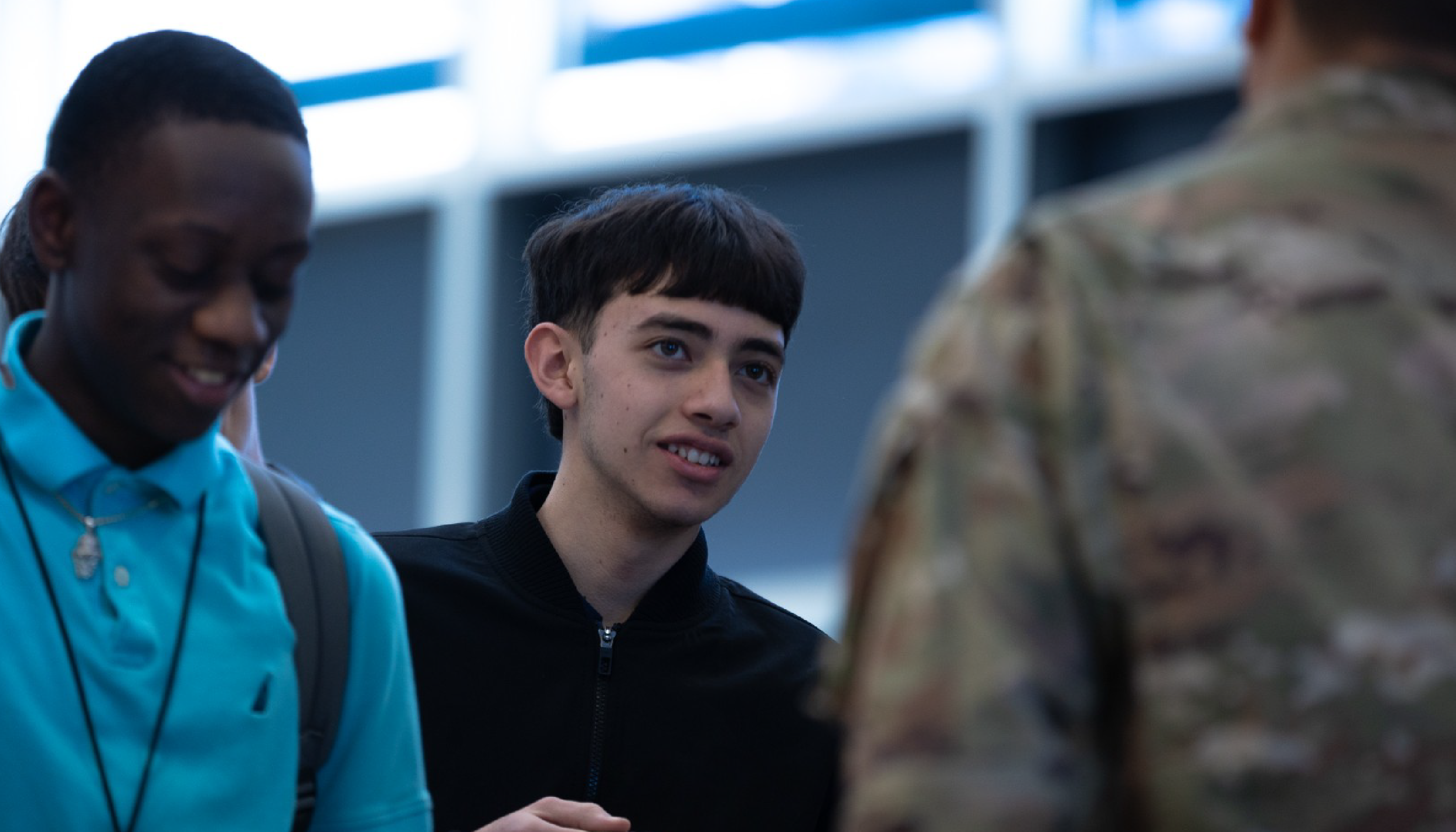 The image shows three young men standing together, with one wearing a turquoise jacket, another in a black jacket, and the third in a camouflage jacket. The background appears to be a city street or urban setting.