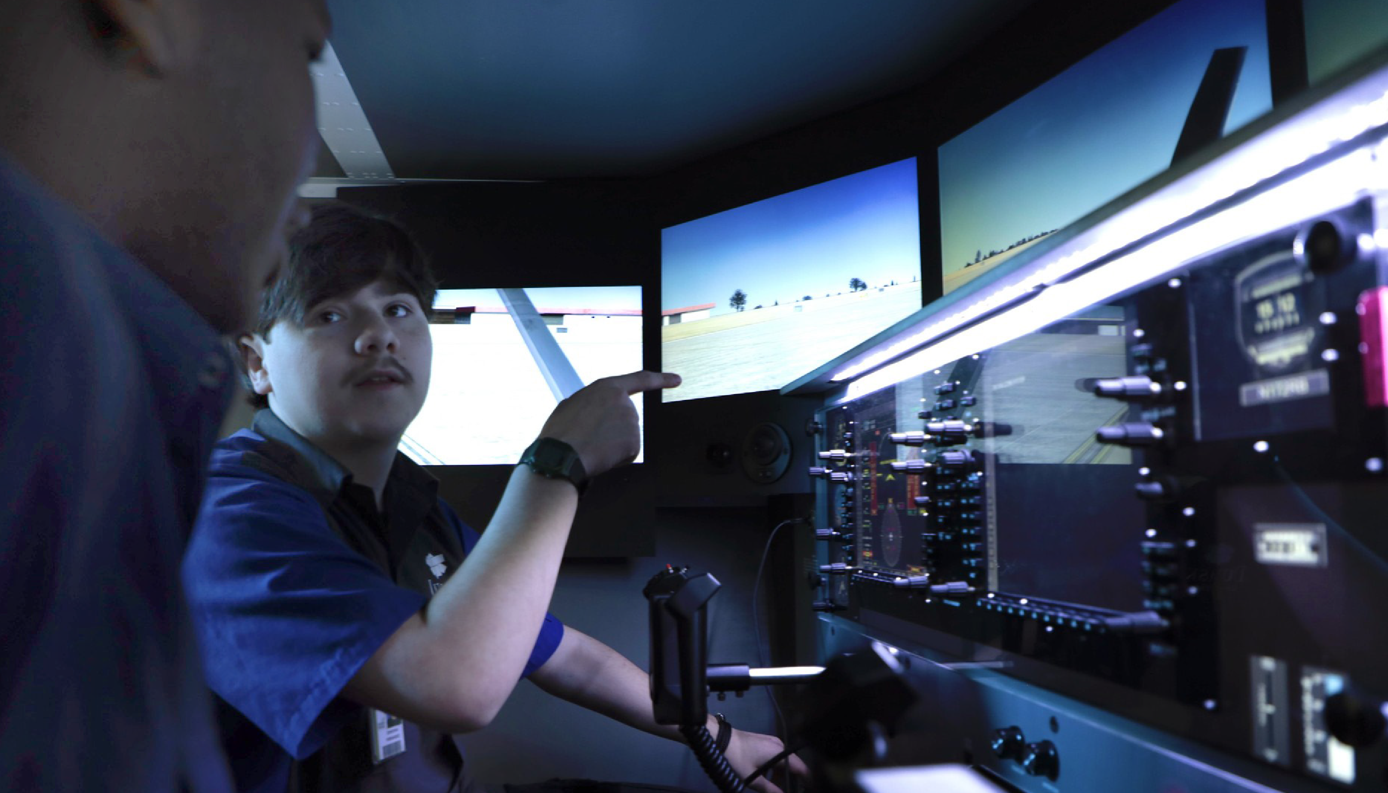 A person in a blue uniform is operating various controls and displays in what appears to be a control room or cockpit setting, with multiple screens and panels visible in the background.