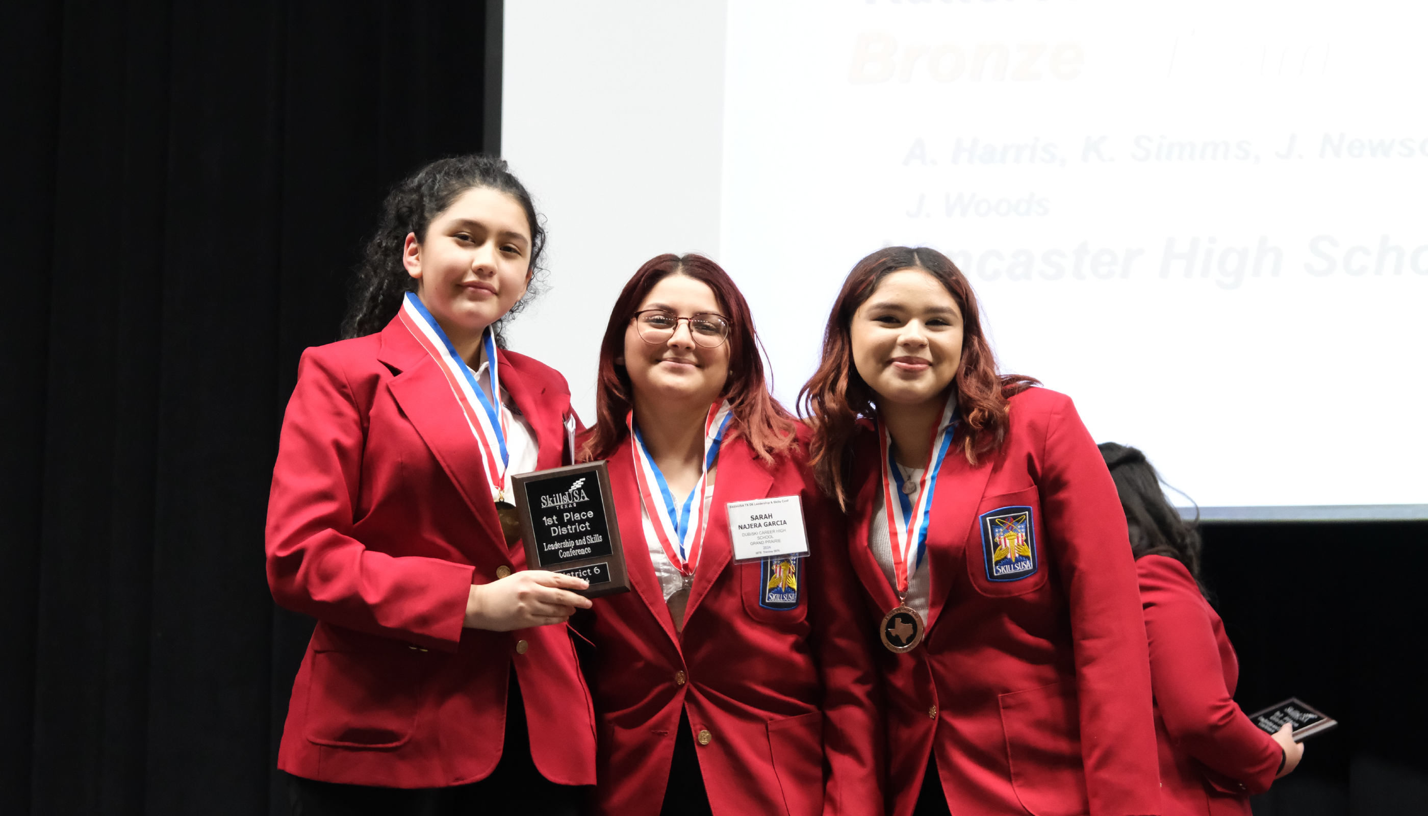 Three young women in red blazers and badges stand together, smiling and holding an award.