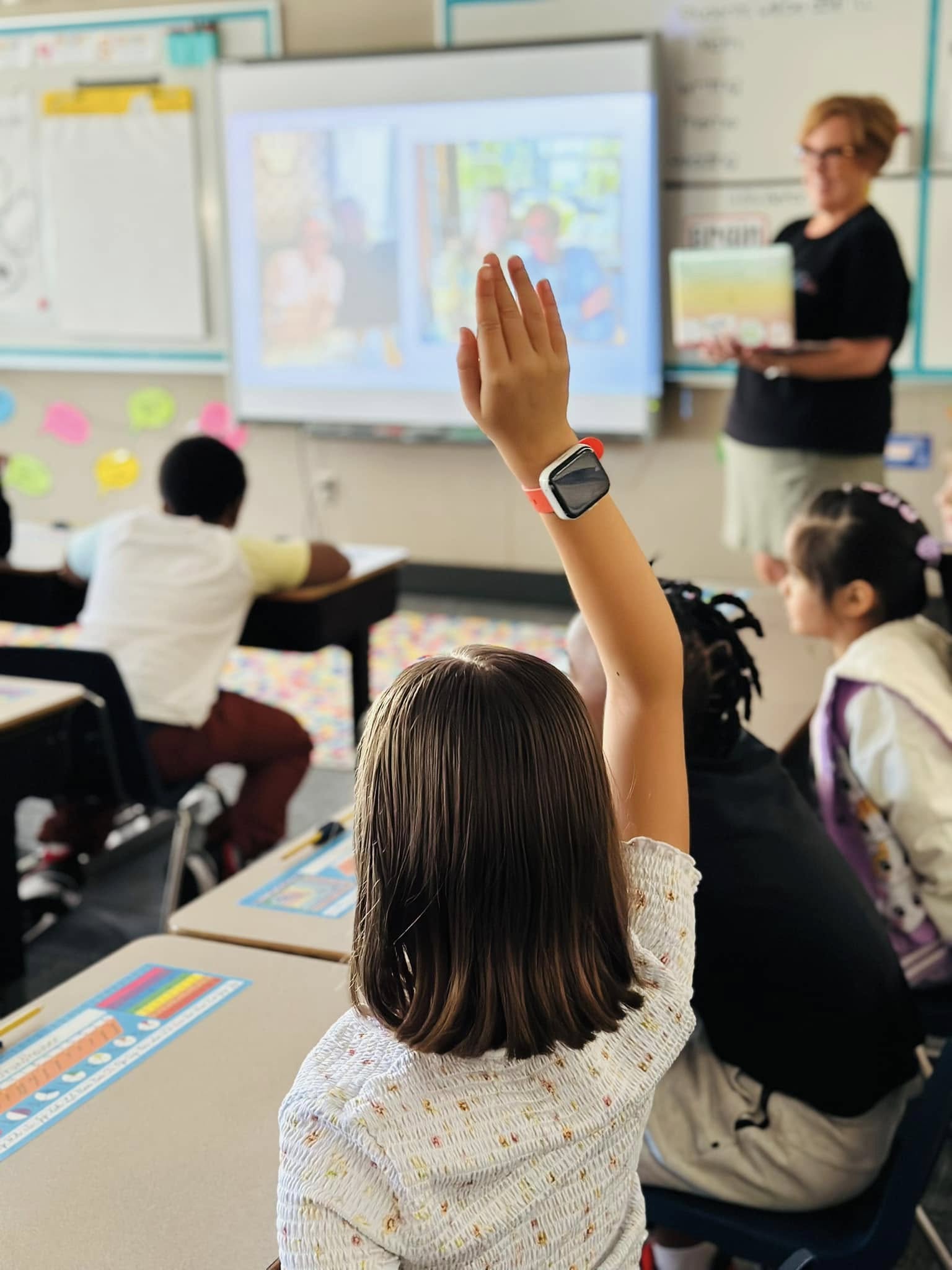A young student with a raised hand in the foreground is in a classroom where a teacher stands near a projector screen in the background.