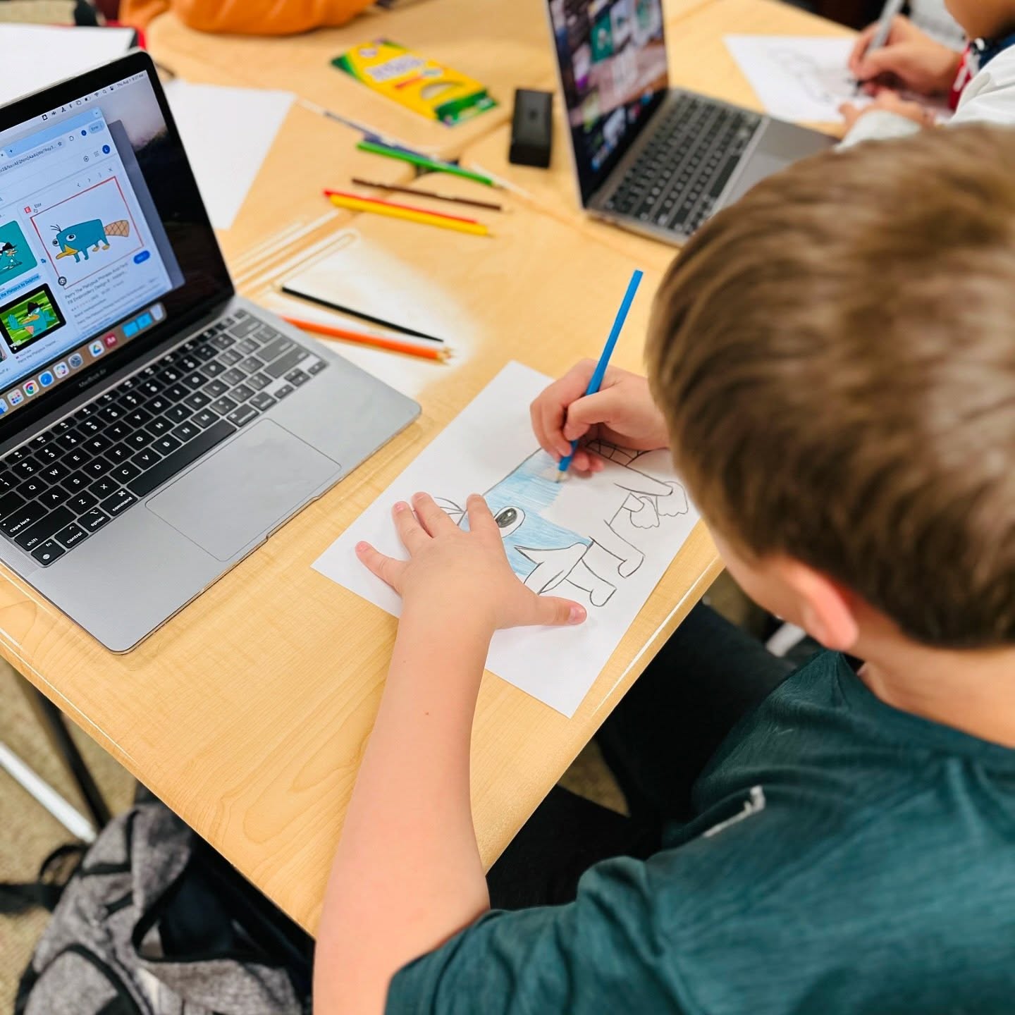 A young boy colors a cartoon dog on paper with a blue crayon, while a laptop displays a similar image in the foreground, and other students work on their own projects in the background.