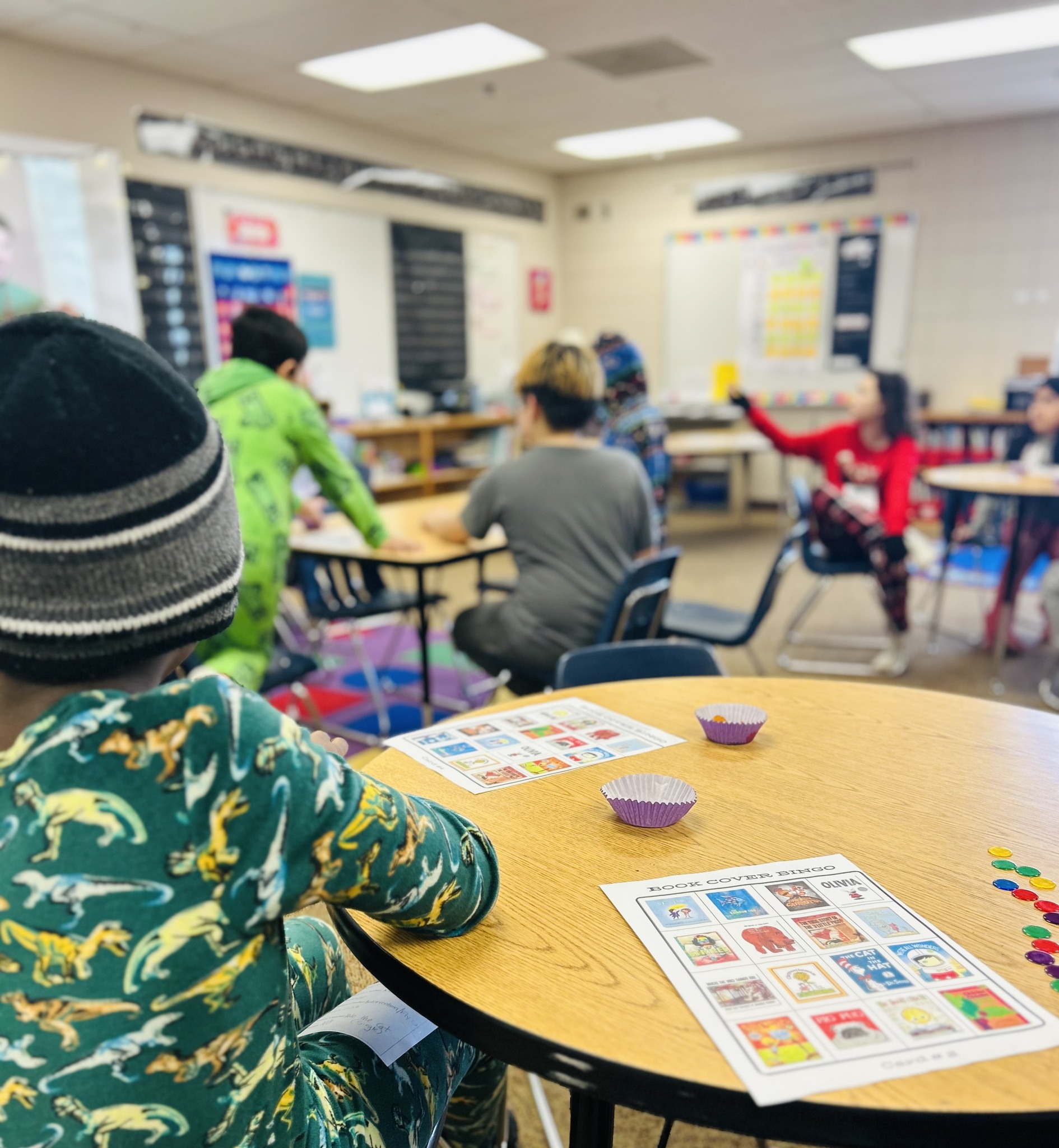 Children in pajamas are gathered around tables with worksheets and colorful tokens in a classroom decorated with student work.