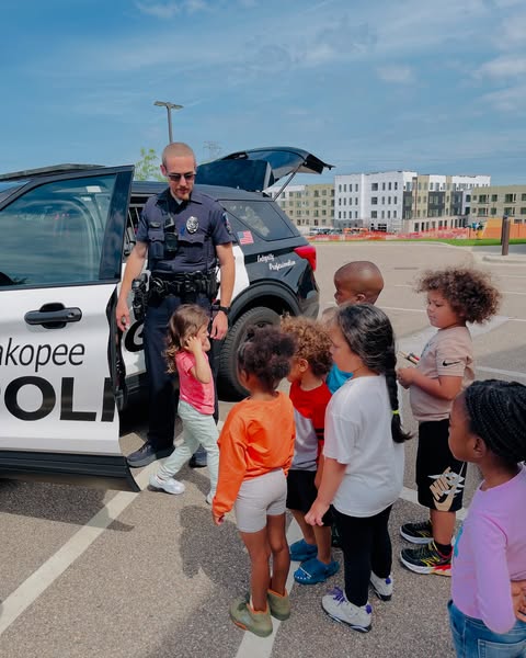 A police officer stands by an open car door, surrounded by a group of young children, with modern buildings visible in the background under a clear sky.
