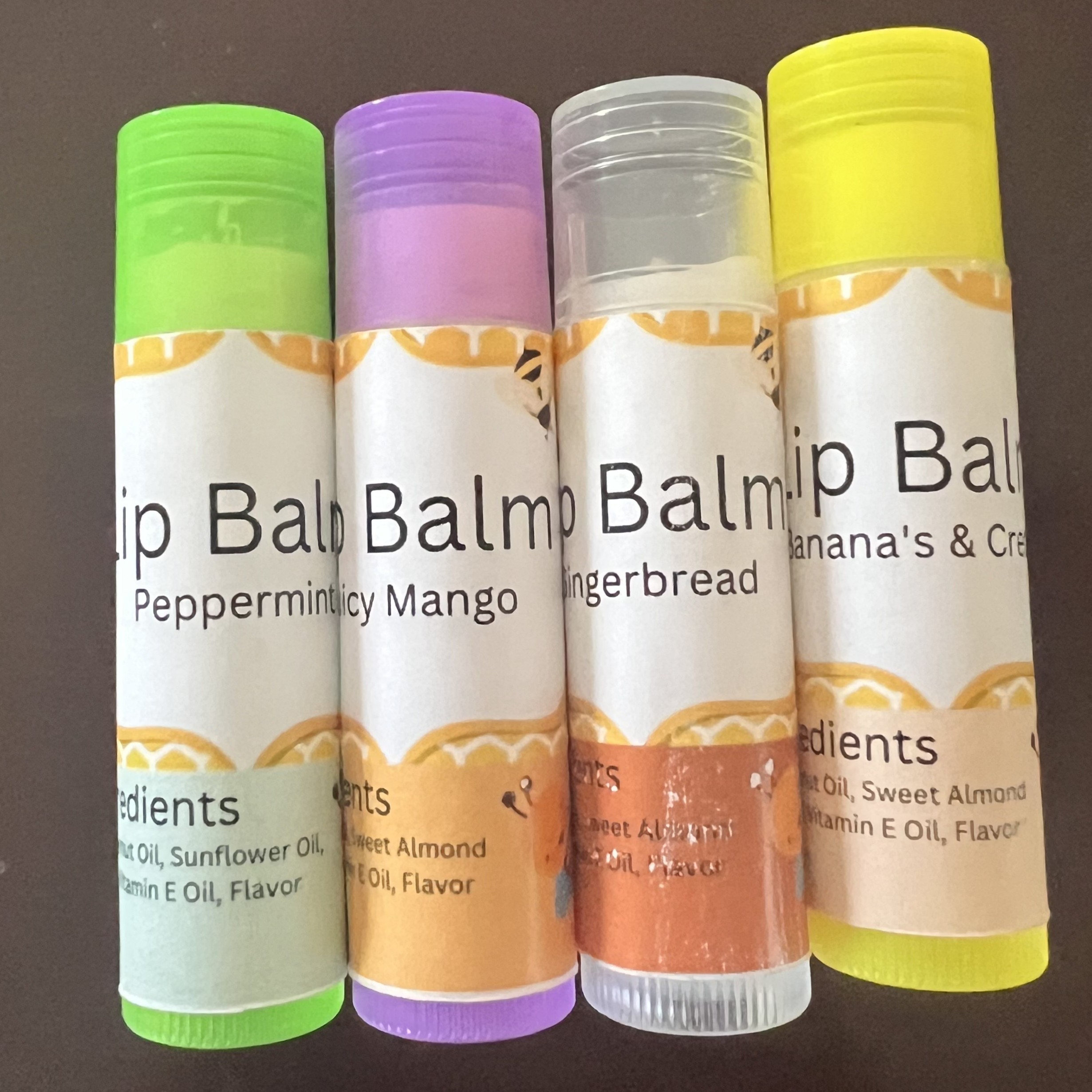 Four colorful lip balm containers with various flavors such as Peppermint Mango, Gingerbread, and Sweet Almond are displayed against a plain brown background.
