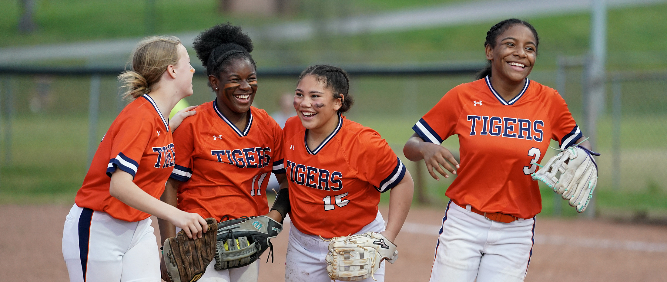 Four softball players in orange jerseys smile and run off the field.