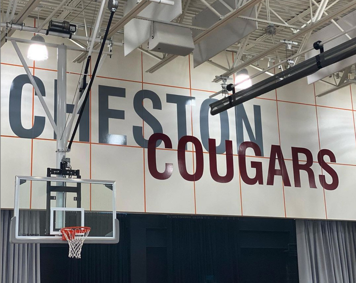 The words Cheston Cougars appear on a white wall above a stage with gray curtains. A basketball hoop appears in the foreground.