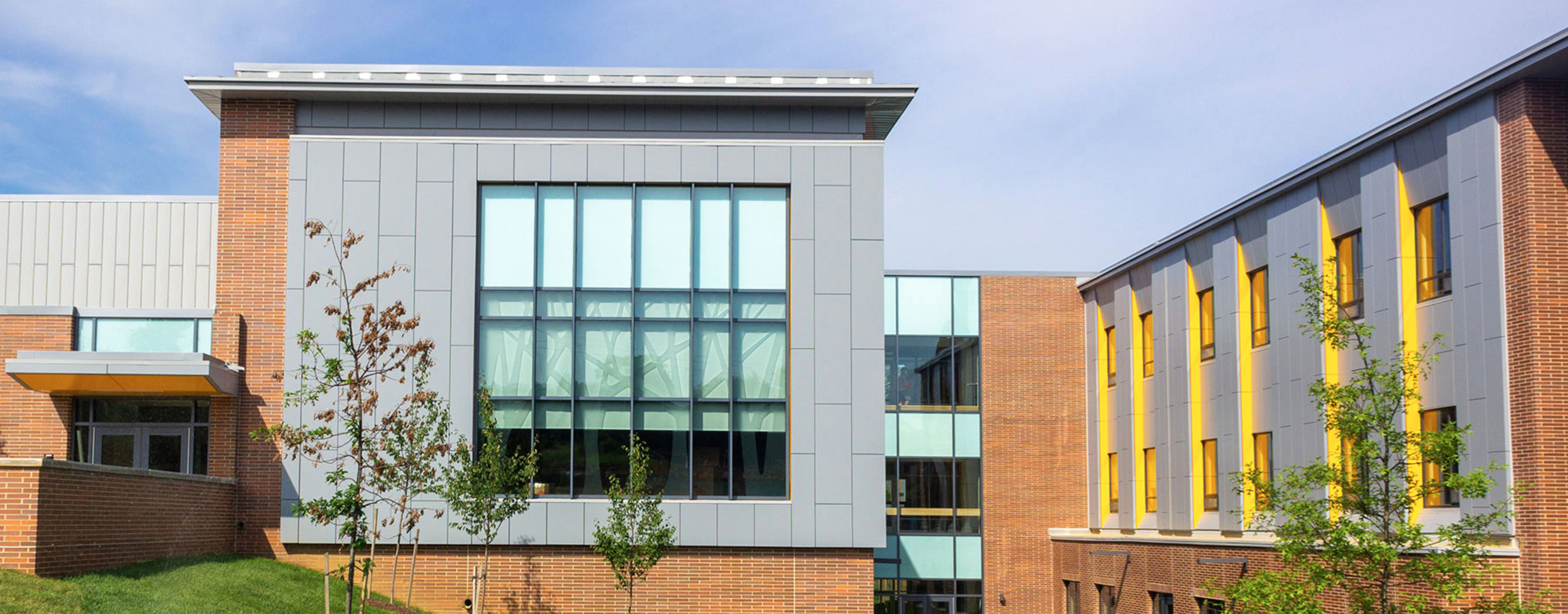 A brick school with yellow and gray trimmings and windows is shown with small trees in front and a blue sky above.