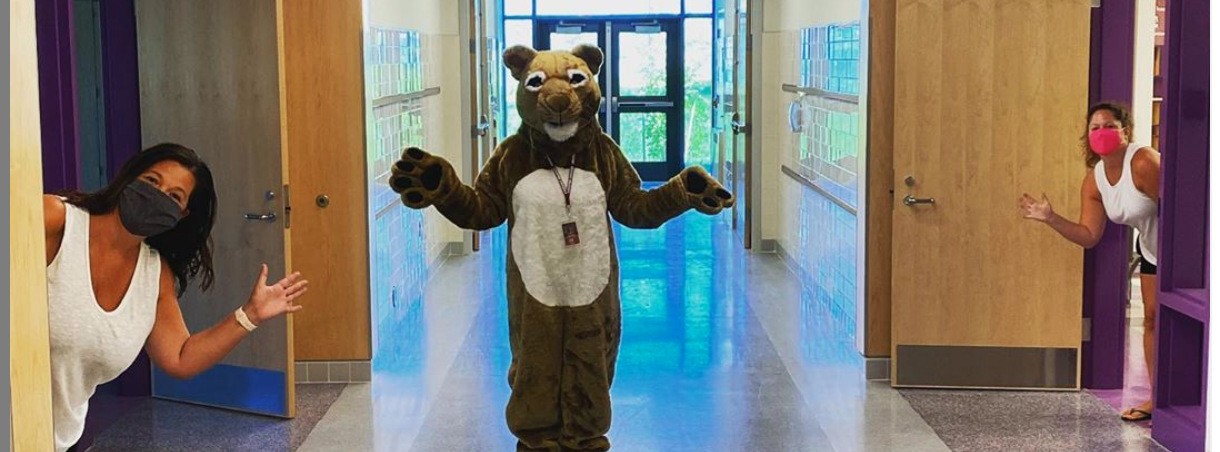 The Cheston Cougar mascot stands in a school hallway with his arms out to the side. Two adult females are leaning in from each side of the hallway and are waving to the camera. 