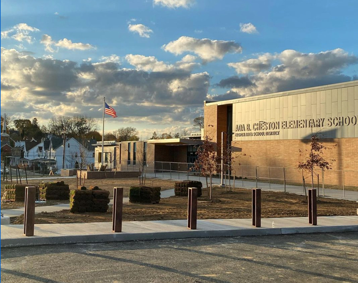 A photo of the front entrance of Cheston Elementary School. A mostly blue sky is filled with puffs of white and gray clouds and an American flag flies in front of the building.
