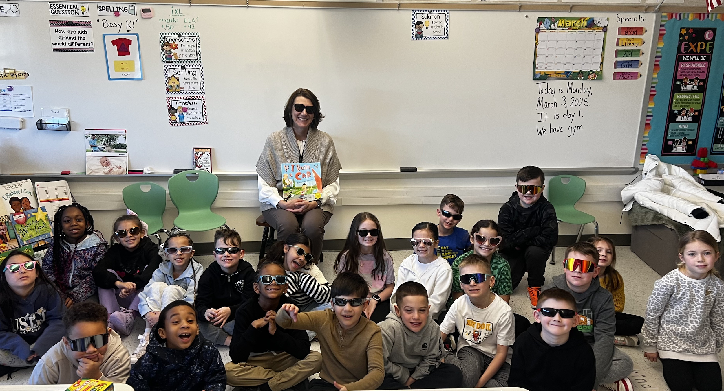 Elementary aged students sit on the floor in a classroom, on a colorful carpet and an adult sitting in a chair with a book all look at the camera and smile.