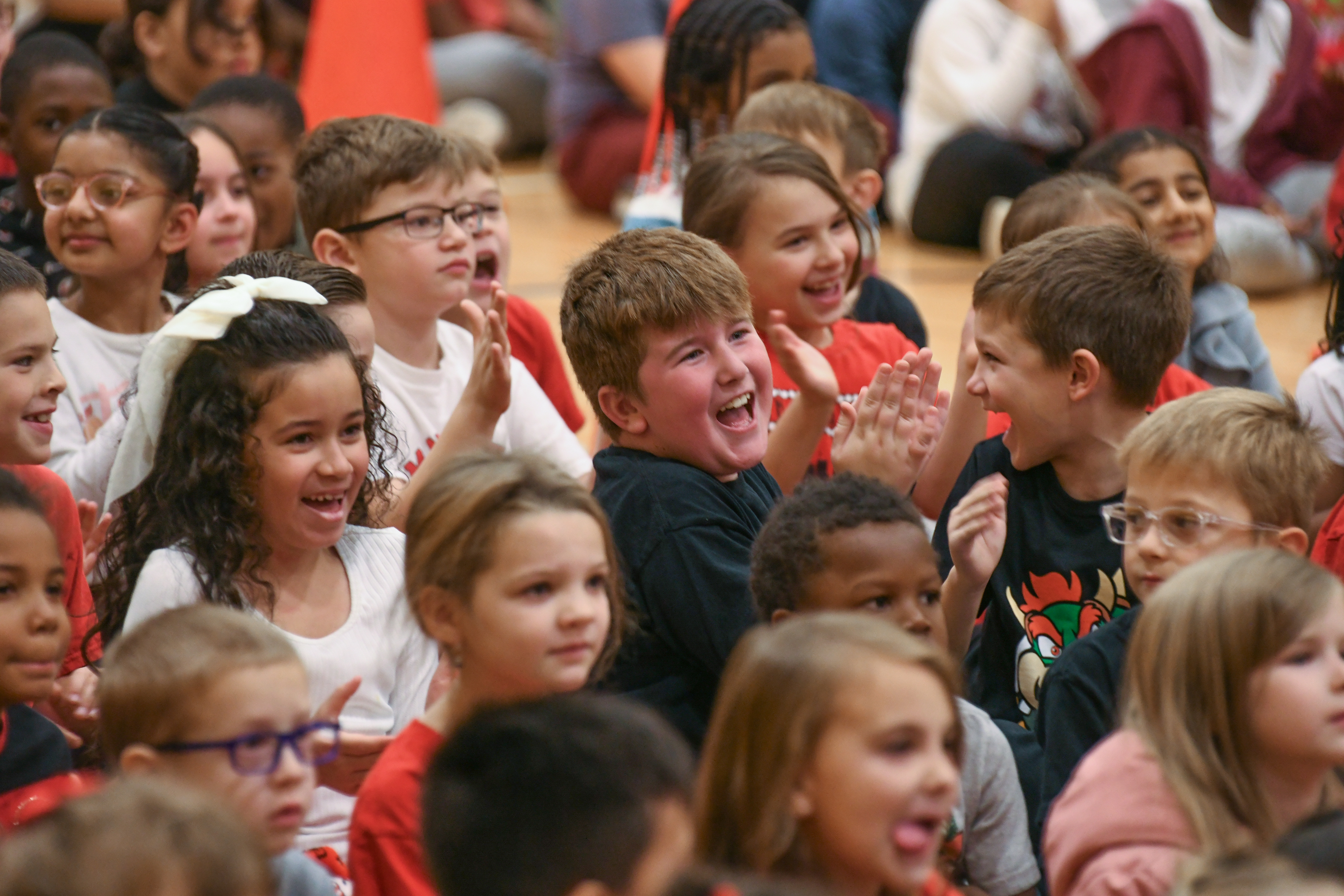 A small group of elementary aged students sit together on a gymnasium floor, smiling and clapping.