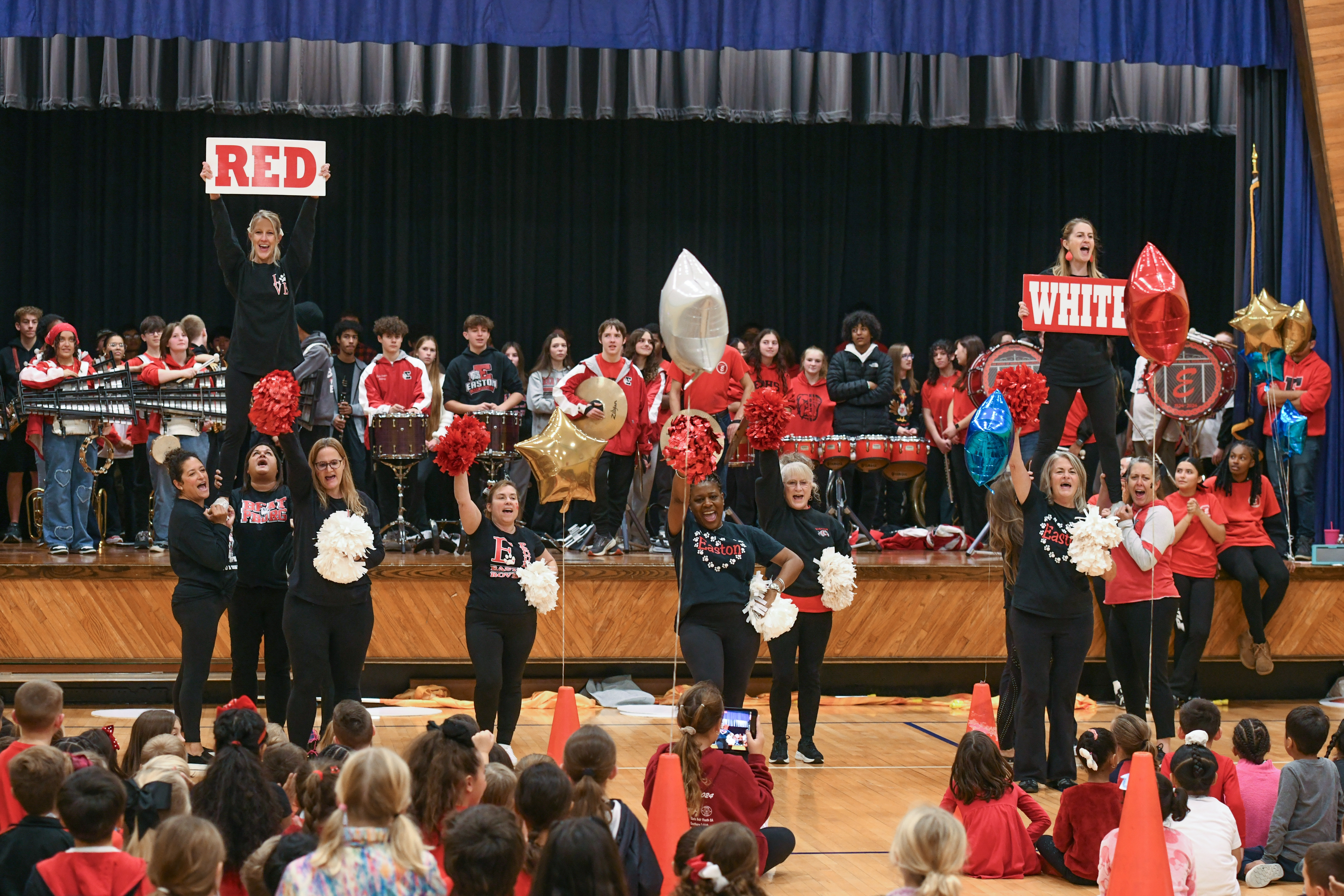 High school aged students playing instruments stand on a stage as a group of adults cheerleaders hold pom-poms and signs in front of them. Elementary aged students sit on the floor facing them.