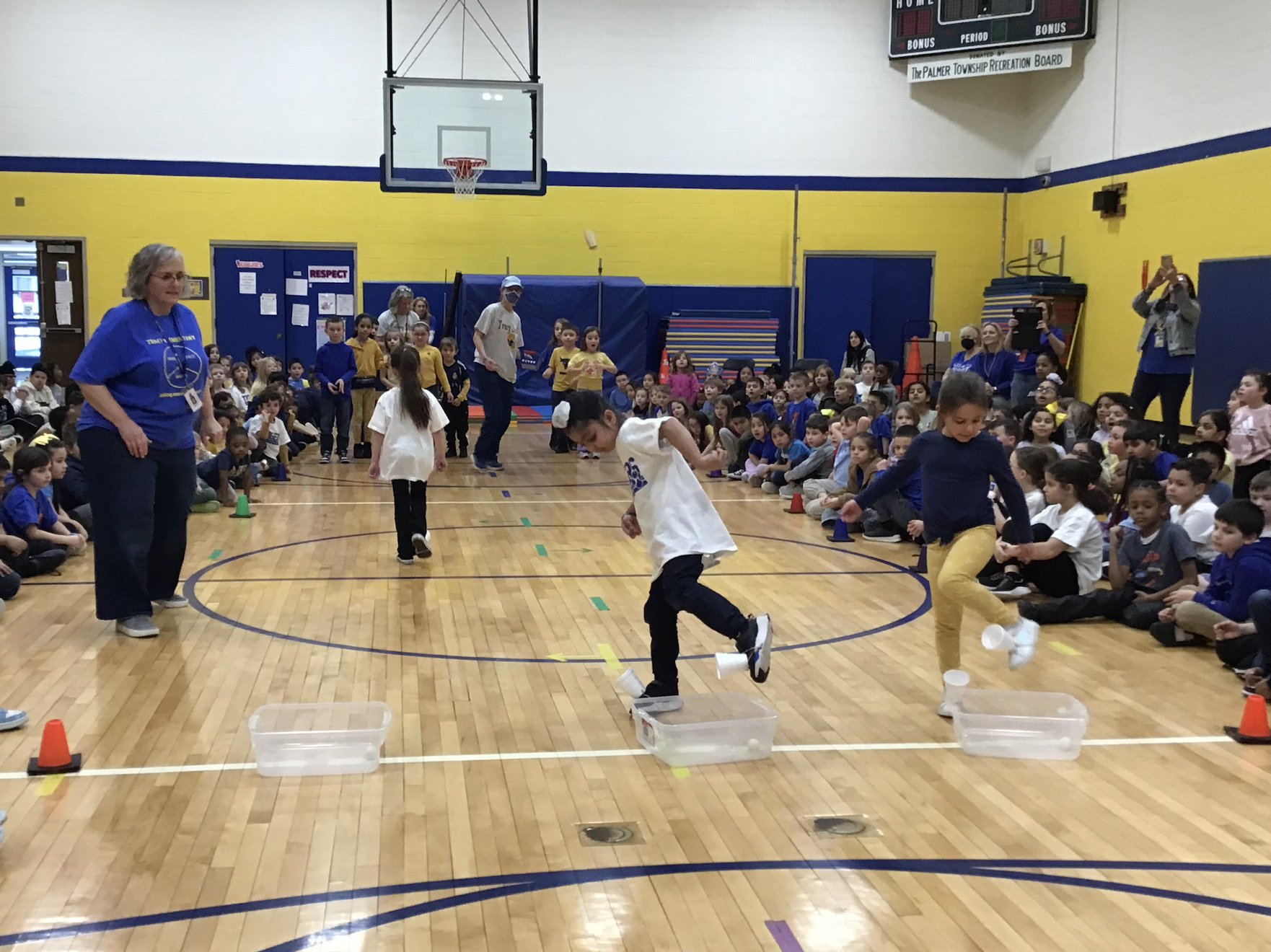 Elementary students sit along two sides of a gymnasium floor as they watch 3 students with cups attached to the tops of their sneakers try to scoop a ping pong ball from a clear plastic container that sits on the floor.