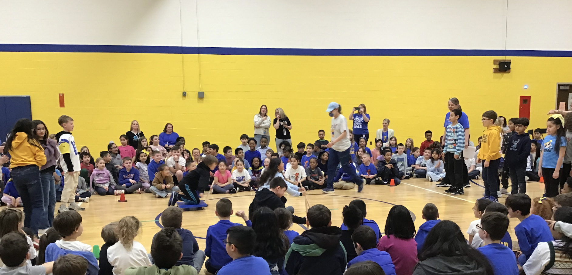 Elementary aged students sit on a gym floor in a square as three students ride scooters across the floor. The gym walls are white on top and yellow on the bottom, with a blue stripe around the middle.