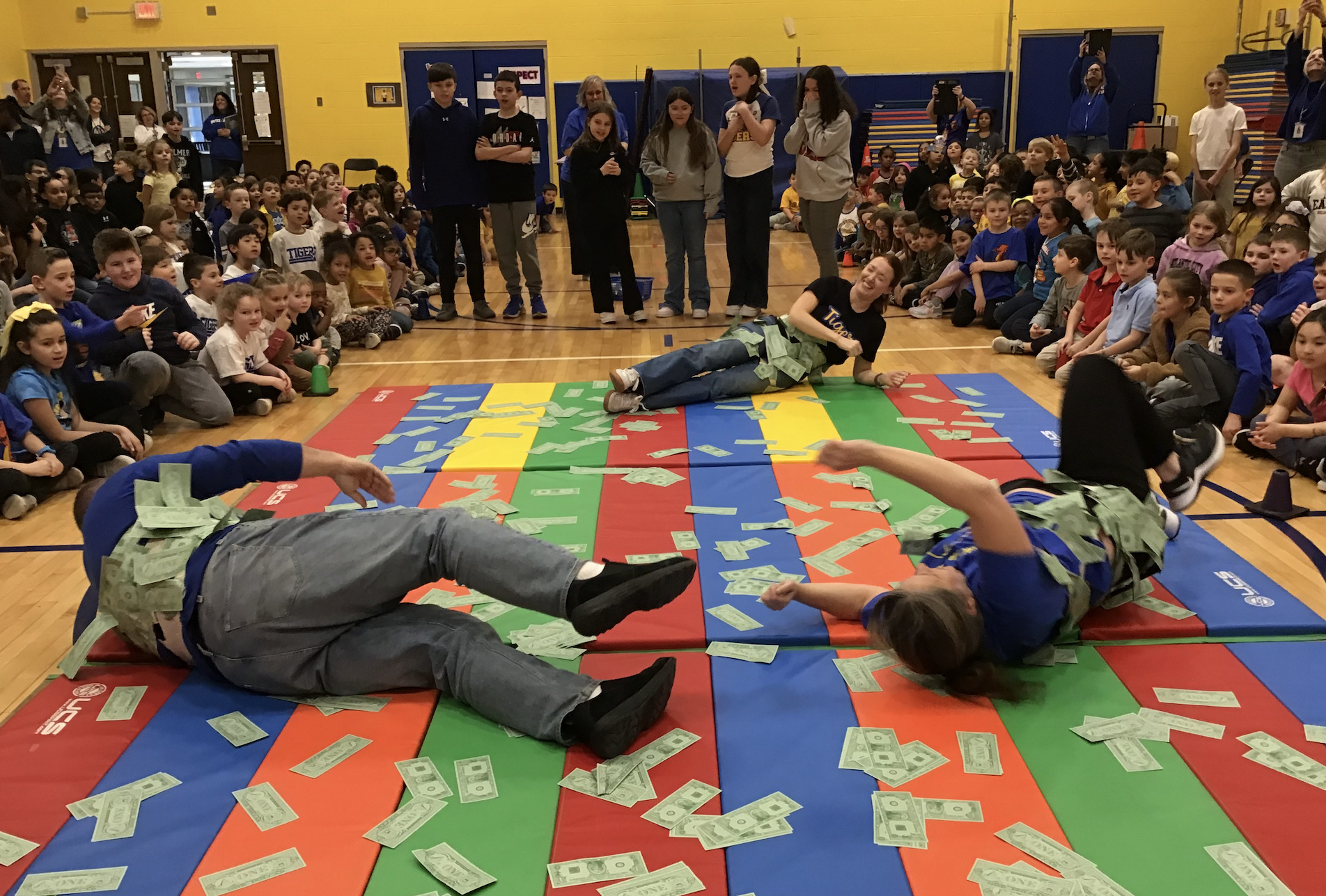Elementary students sit along two sides of a gymnasium floor as they watch 3 adults roll on colorful mats with paper money laying on them. The adults are rolling on the money to get the money stuck to their clothing.