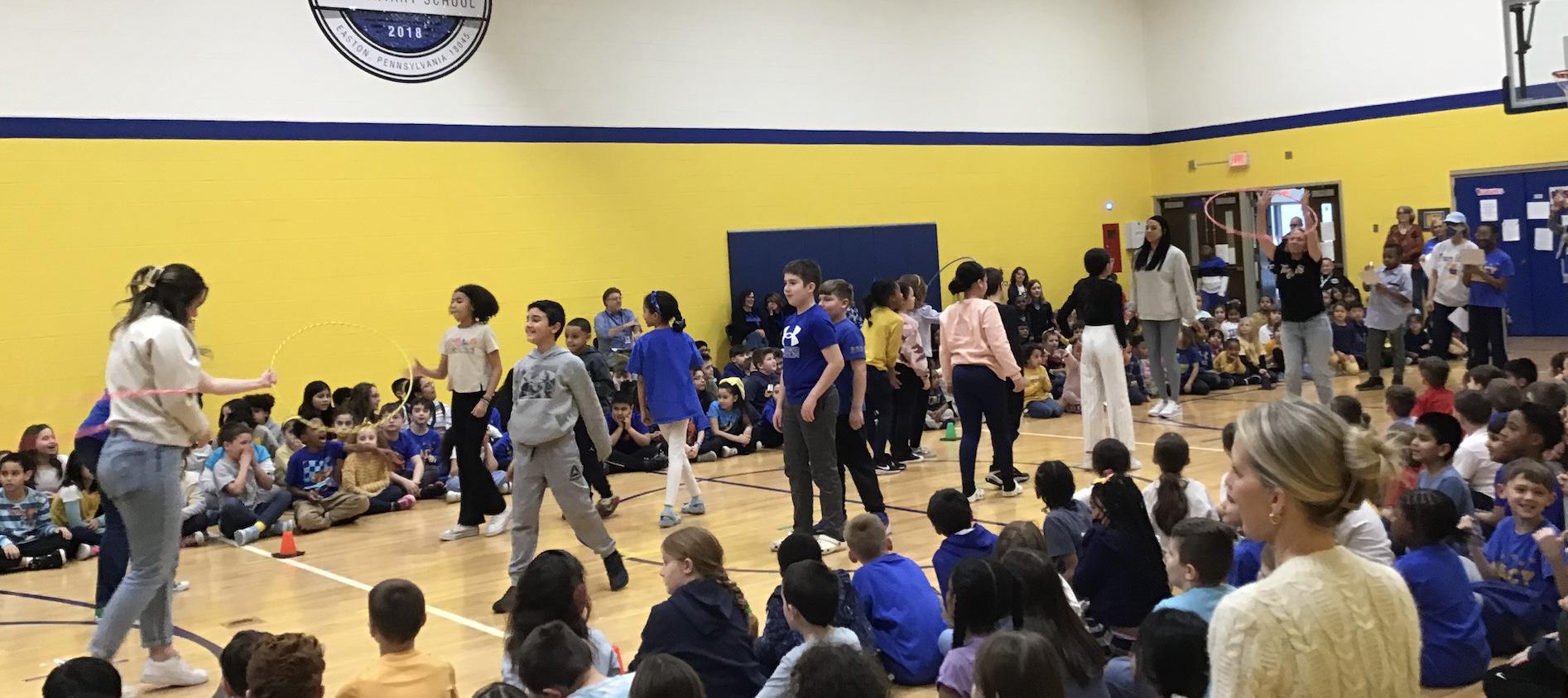 Elementary students sit along two sides of a gymnasium floor as they watch two rows of students who are standing race to pass a hula hoop down their row.
