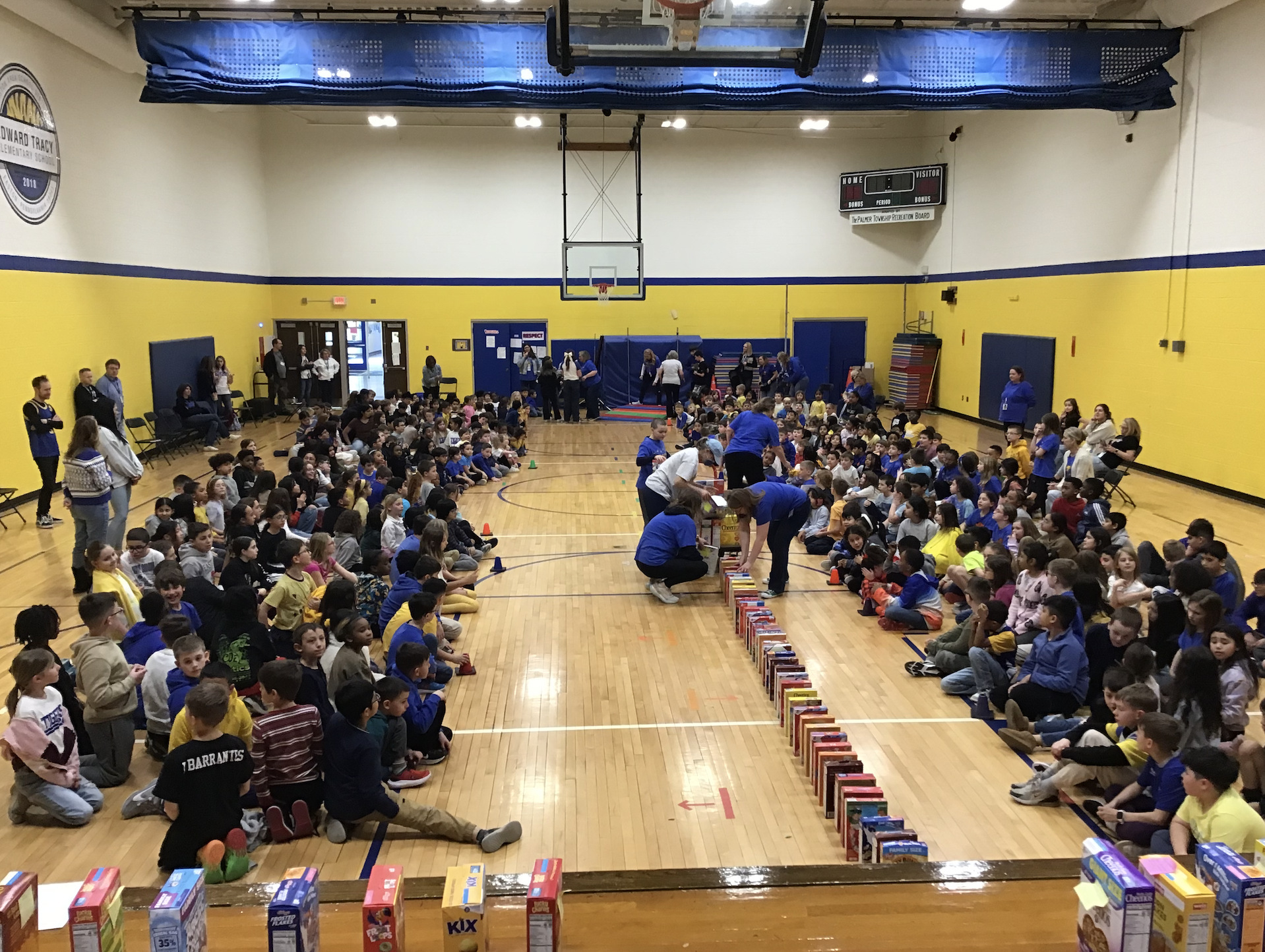 Elementary students sit along two sides of a gymnasium floor as they watch several adults line cereal boxes down the center of the gym floor, set up like dominoes.