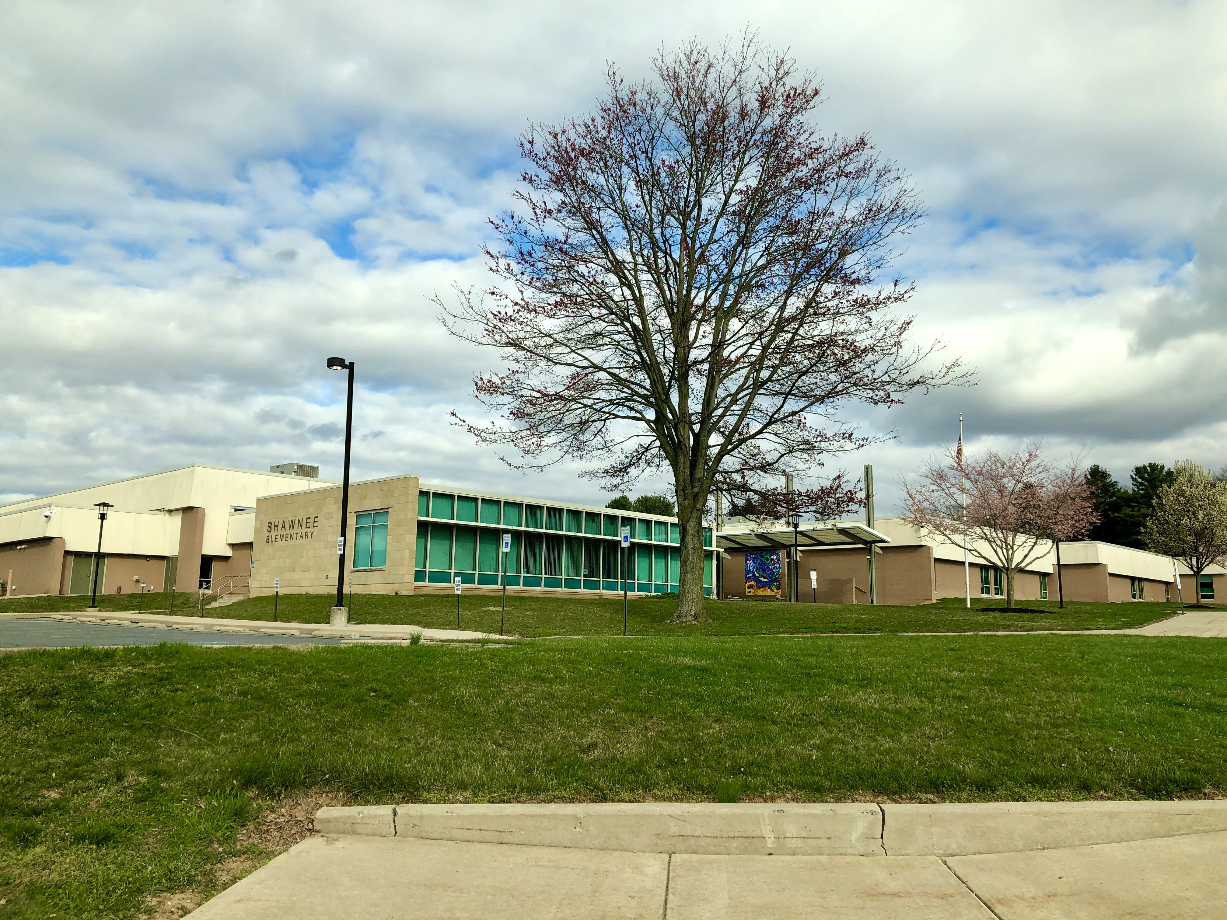 A photo of Shawnee Elementary School, with tan brick, a large tree in the foreground has lost its leaves and sits in a green lawn in front of the school.