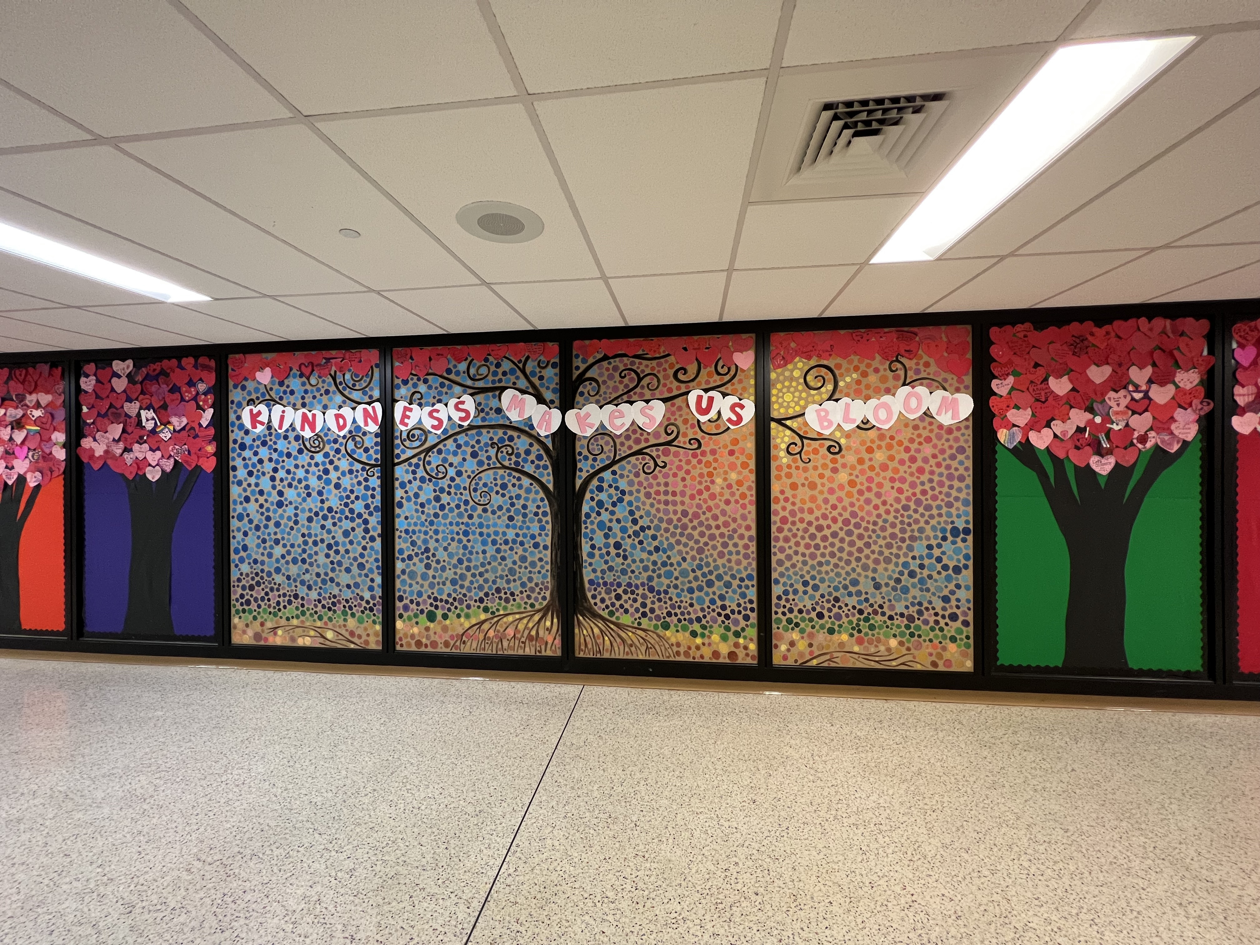 A large interior wall displays several wall panels, four of which create a large tree with colorful dots creating a sky and sun in the background. White paper hearts span the panels and say "Kindness make us bloom."