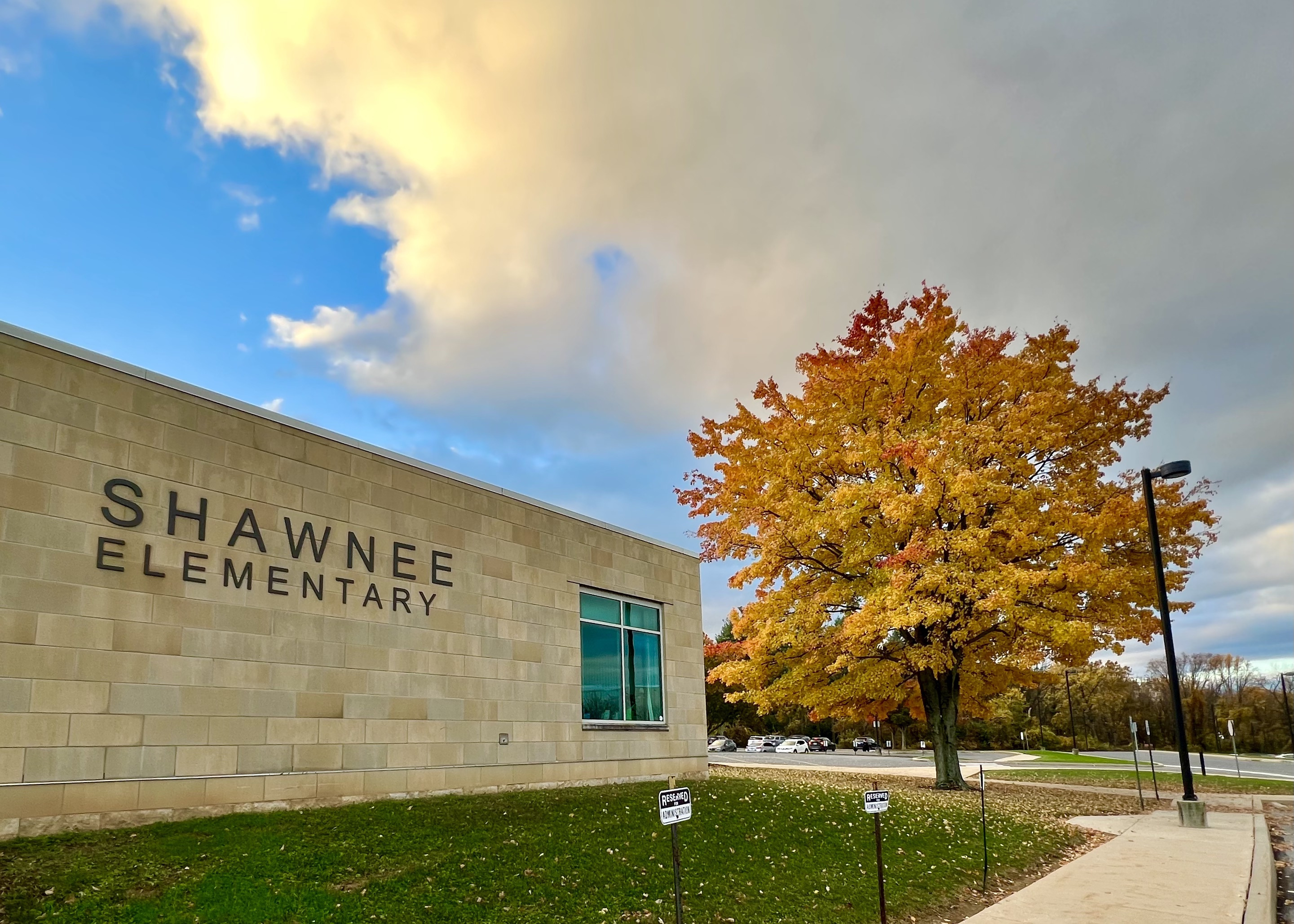 A tan brick school building with the words Shawnee Elementary is shown with a blue sky and clouds. A tree in fall foliage colors sits next to the building in a grassy area.